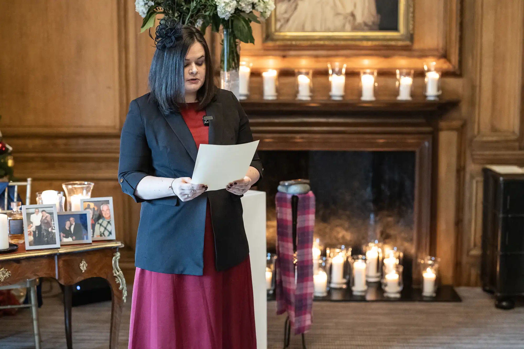 A woman in a black blazer and red dress reads from a paper in a room with lit candles, framed photographs, and floral arrangements.