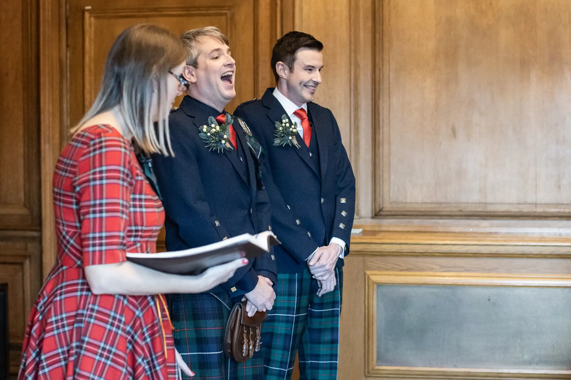 Two grooms in matching plaid kilts and blazers stand next to a woman reading from a book in a wood-paneled room. One groom is laughing heartily.