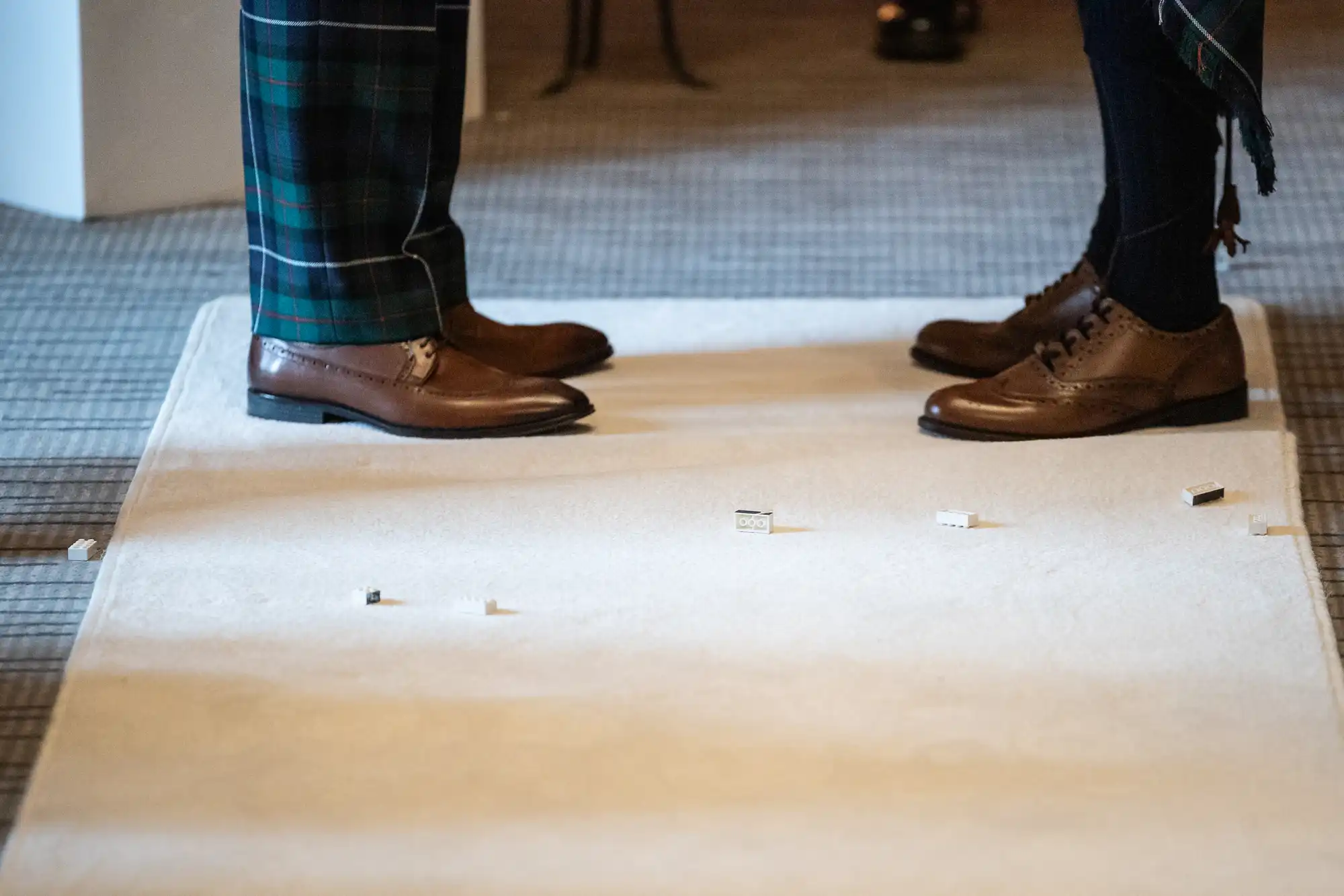 Two individuals wearing brown shoes are standing on a beige carpet, with small scattered items on the carpet near their feet.