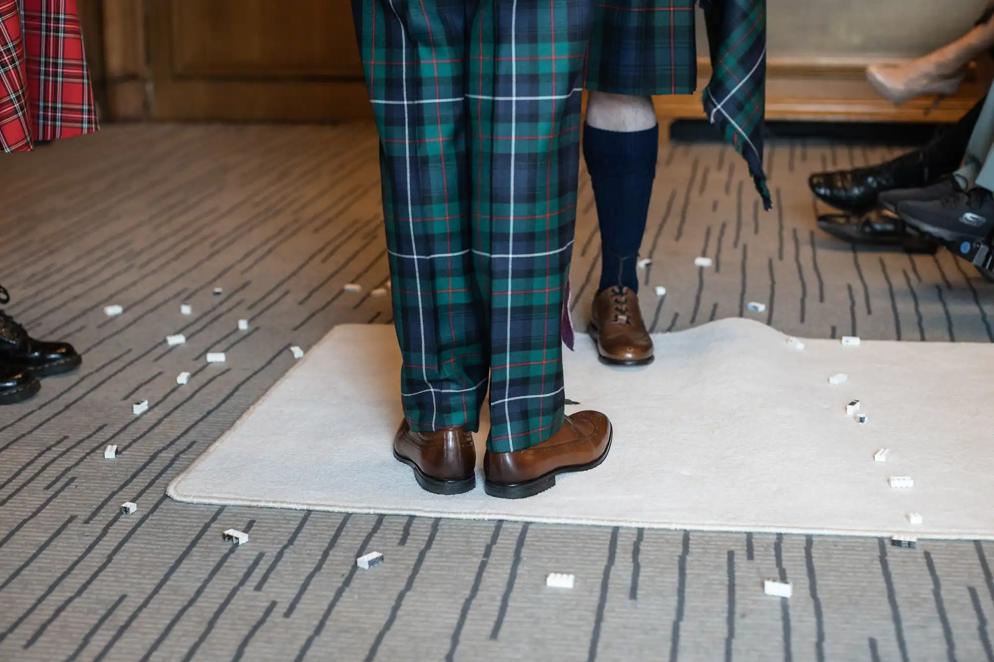 Two people wearing green tartan kilts and brown shoes stand on a white rug scattered with small white pieces, on a striped gray carpet.