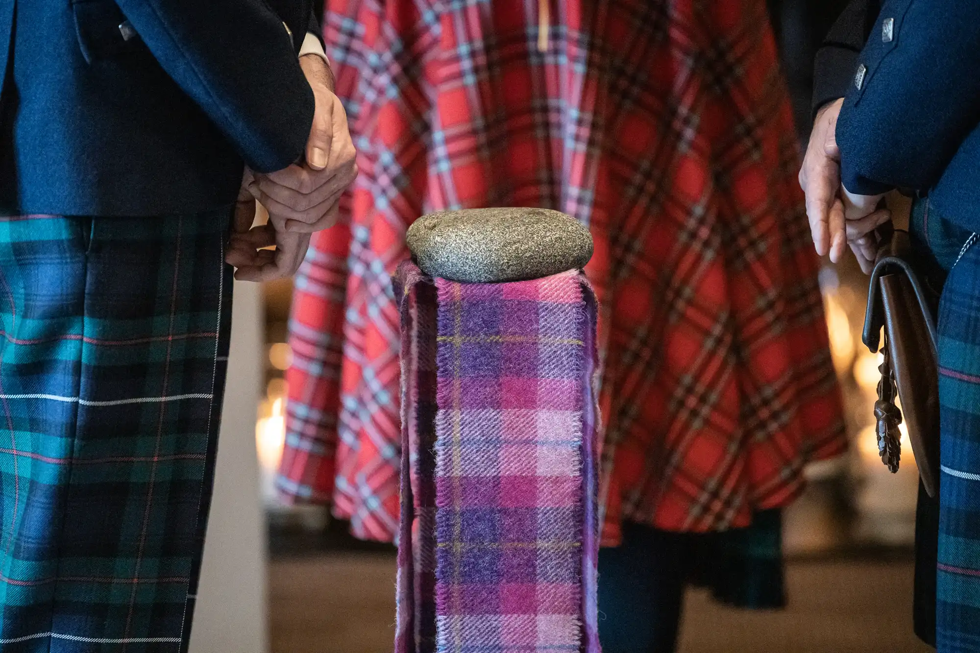 Three people in tartan clothing stand around a stone placed on a fabric-covered pedestal.