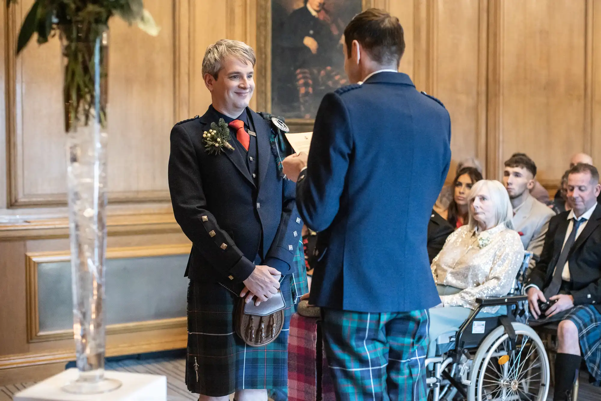 Two men in tartan attire stand facing each other during a ceremony in a wood-paneled room, with guests, including one in a wheelchair, seated and watching.