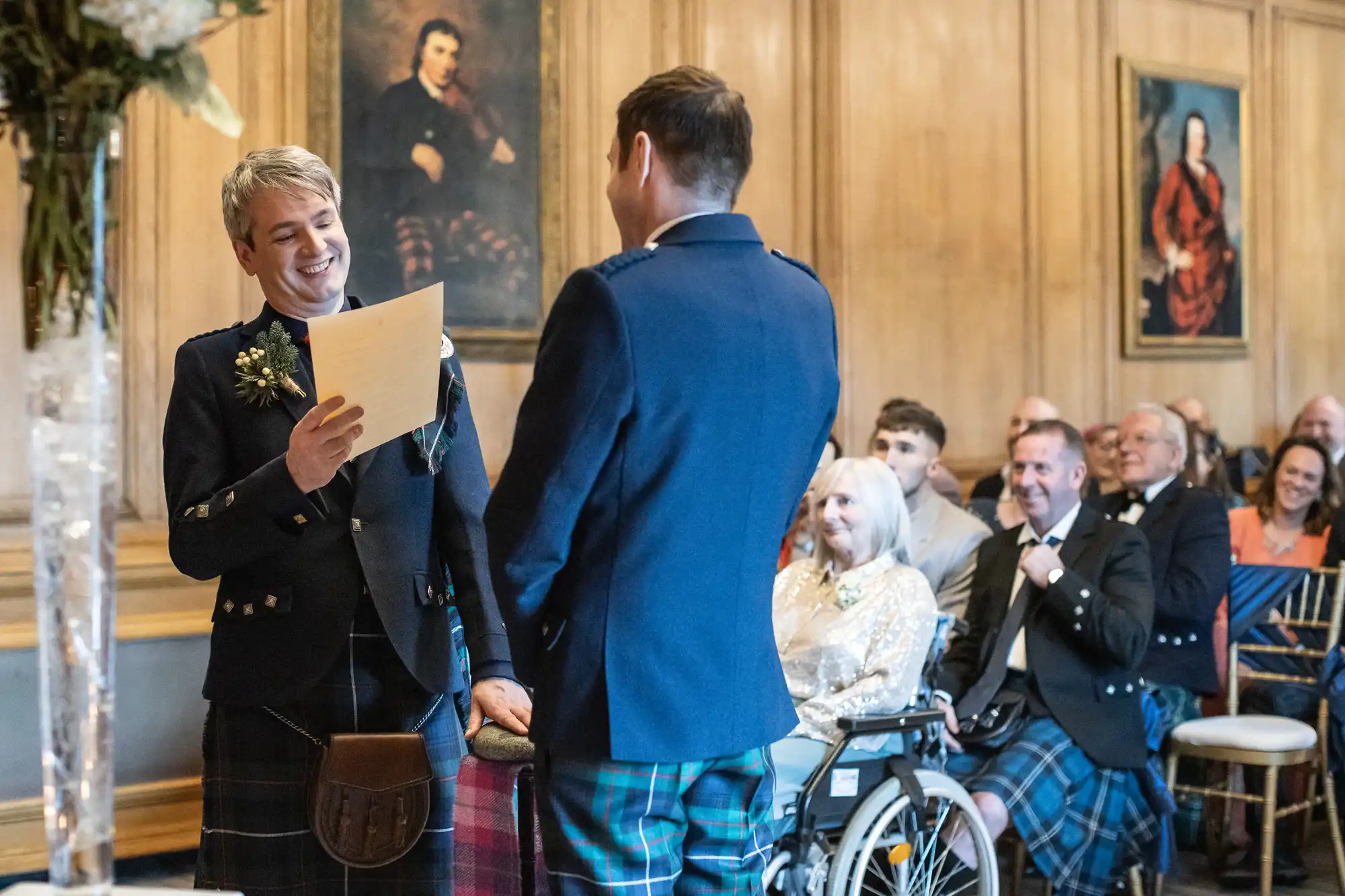 Two men in kilts face each other, one reading from a paper, in a wood-paneled room with people seated and watching, including an elderly woman in a wheelchair.