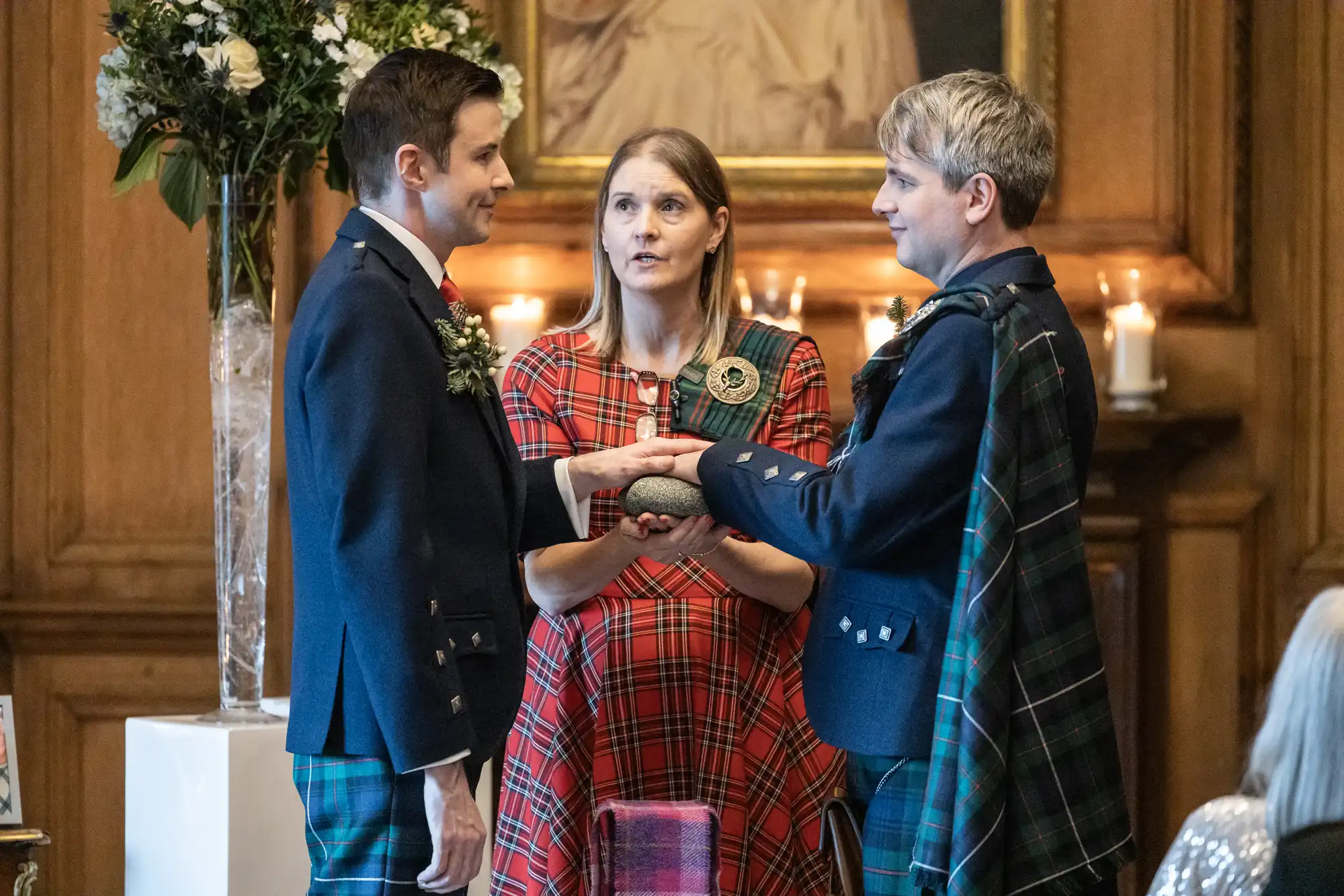 Two grooms in matching tartan attire hold hands during a wedding ceremony officiated by a woman in a red tartan dress, with candles and floral decorations in the background.