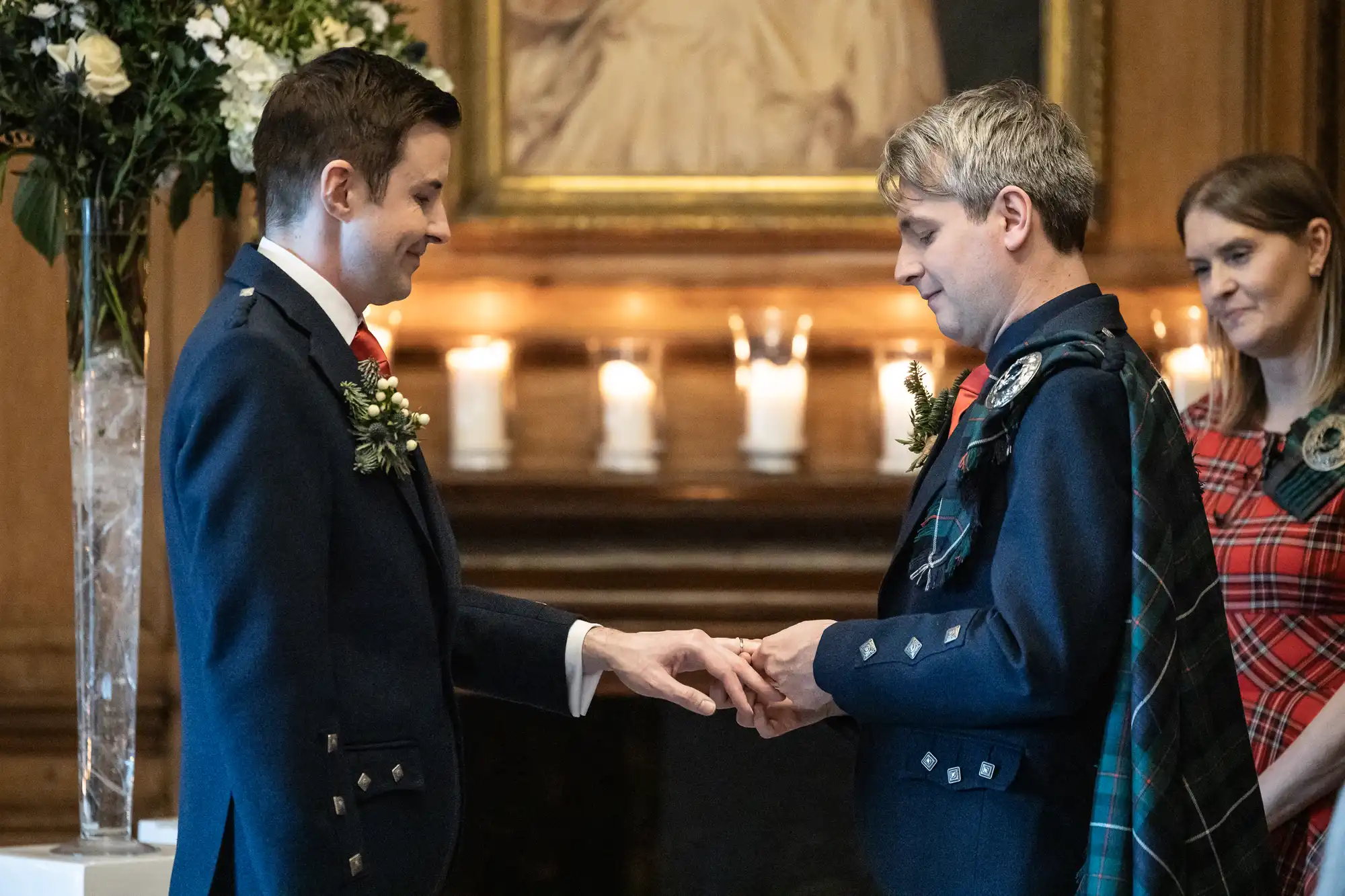 Two men in formal attire exchange rings in a wedding ceremony. A woman in a plaid dress stands in the background, and candles are lit on the mantle behind them.