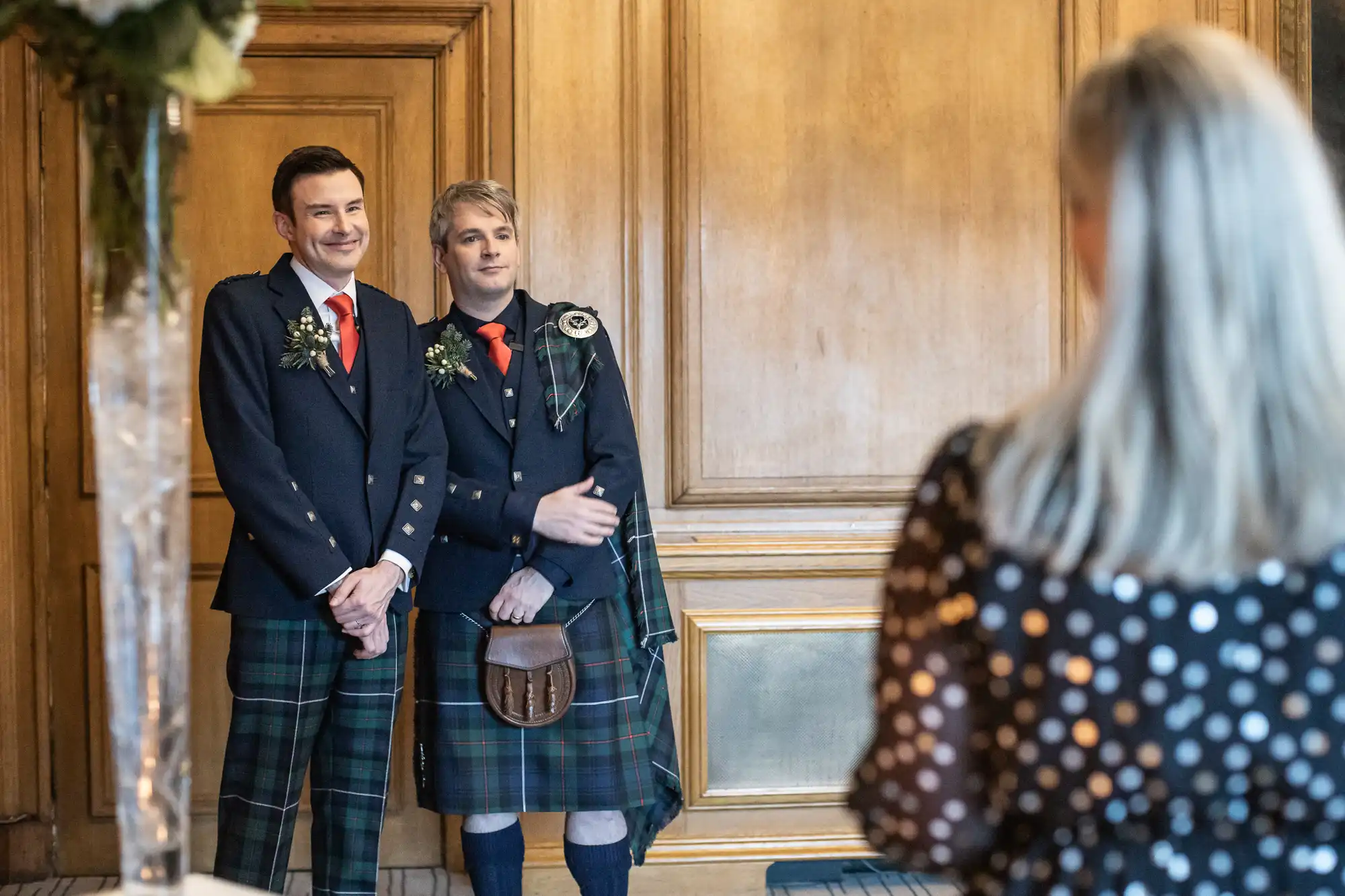 Two men dressed in traditional Scottish kilts and jackets stand together, smiling and holding hands, as they face a woman taking their photo.