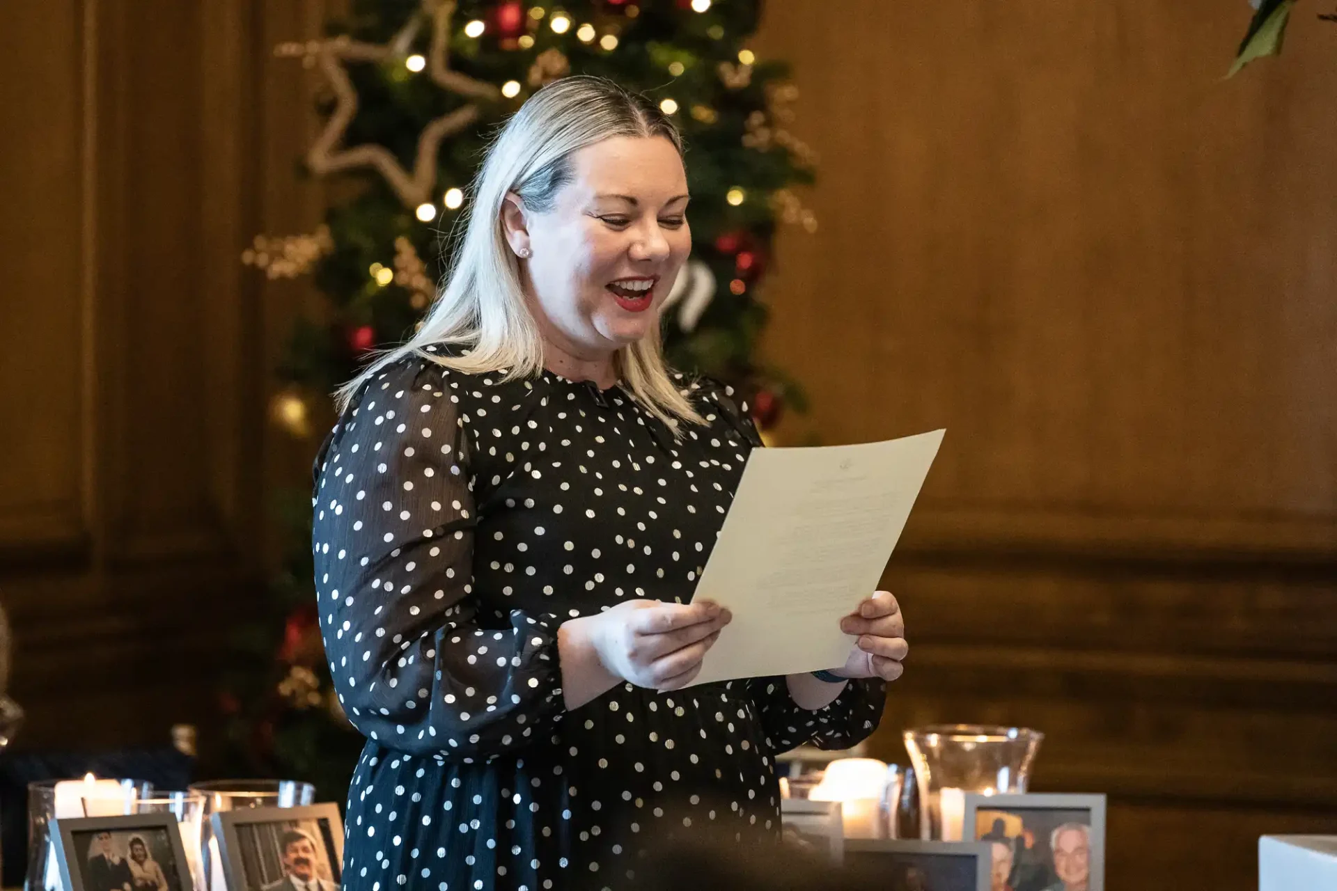 A woman in a black polka dot dress reads a document, smiling, in front of a decorated Christmas tree.