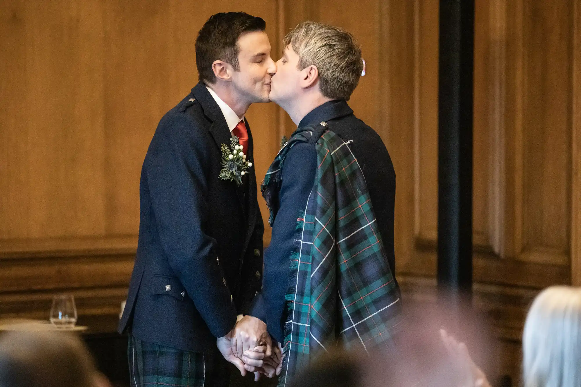 Two men in traditional Scottish attire share a kiss while holding hands in what appears to be a formal event or ceremony in a wooden-paneled room.