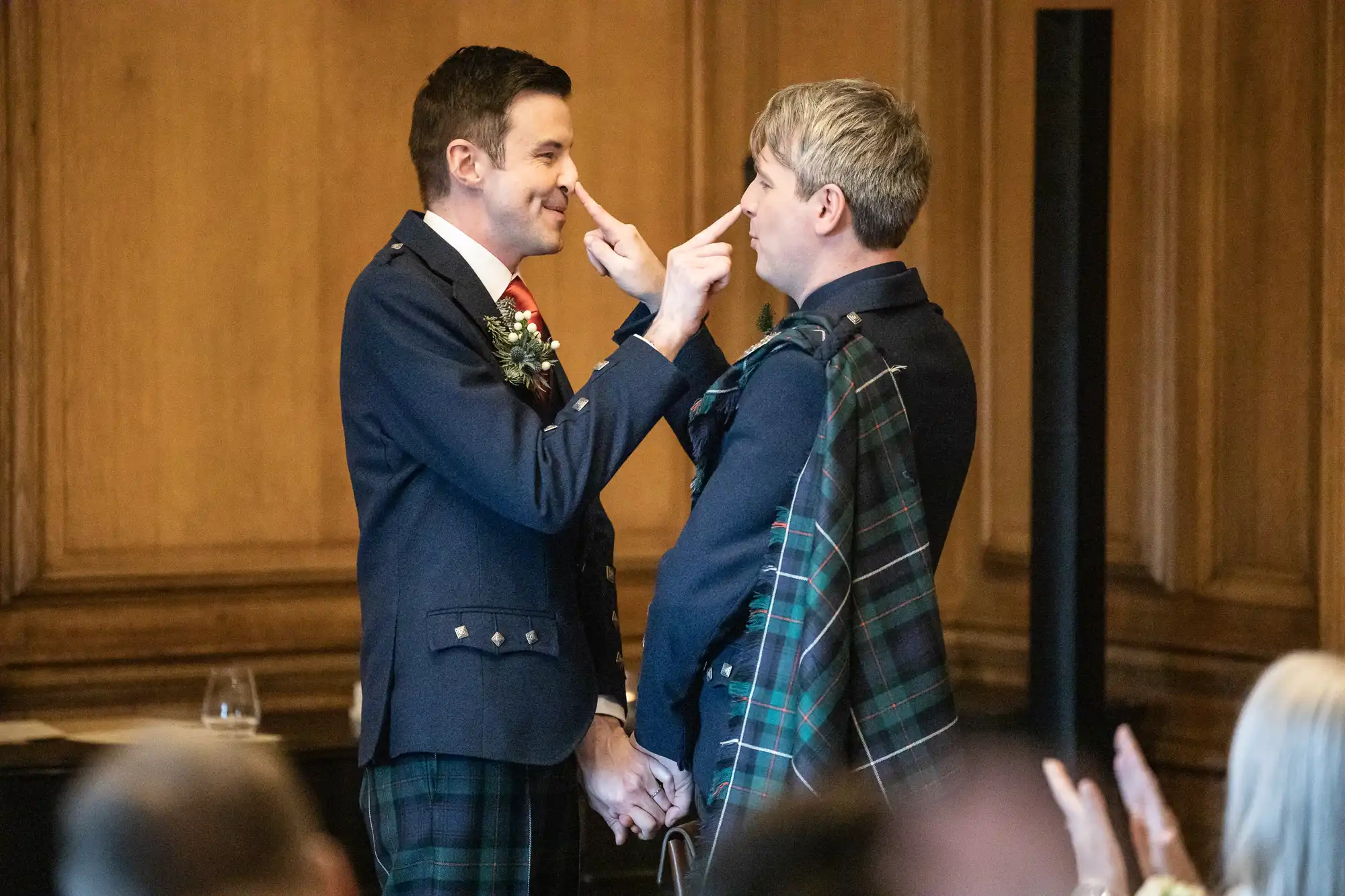 Two men in kilts, dressed in formal attire, stand facing each other in a wood-paneled room. One man touches the other’s nose with his finger while holding hands. People sit watching in the foreground.