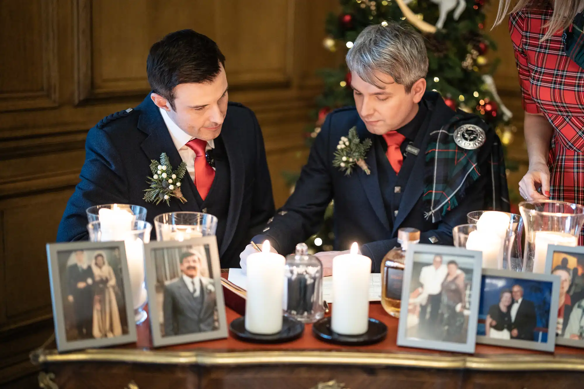 Two men in formal attire sign a document at a candle-lit table adorned with photos and flowers, with a Christmas tree in the background.