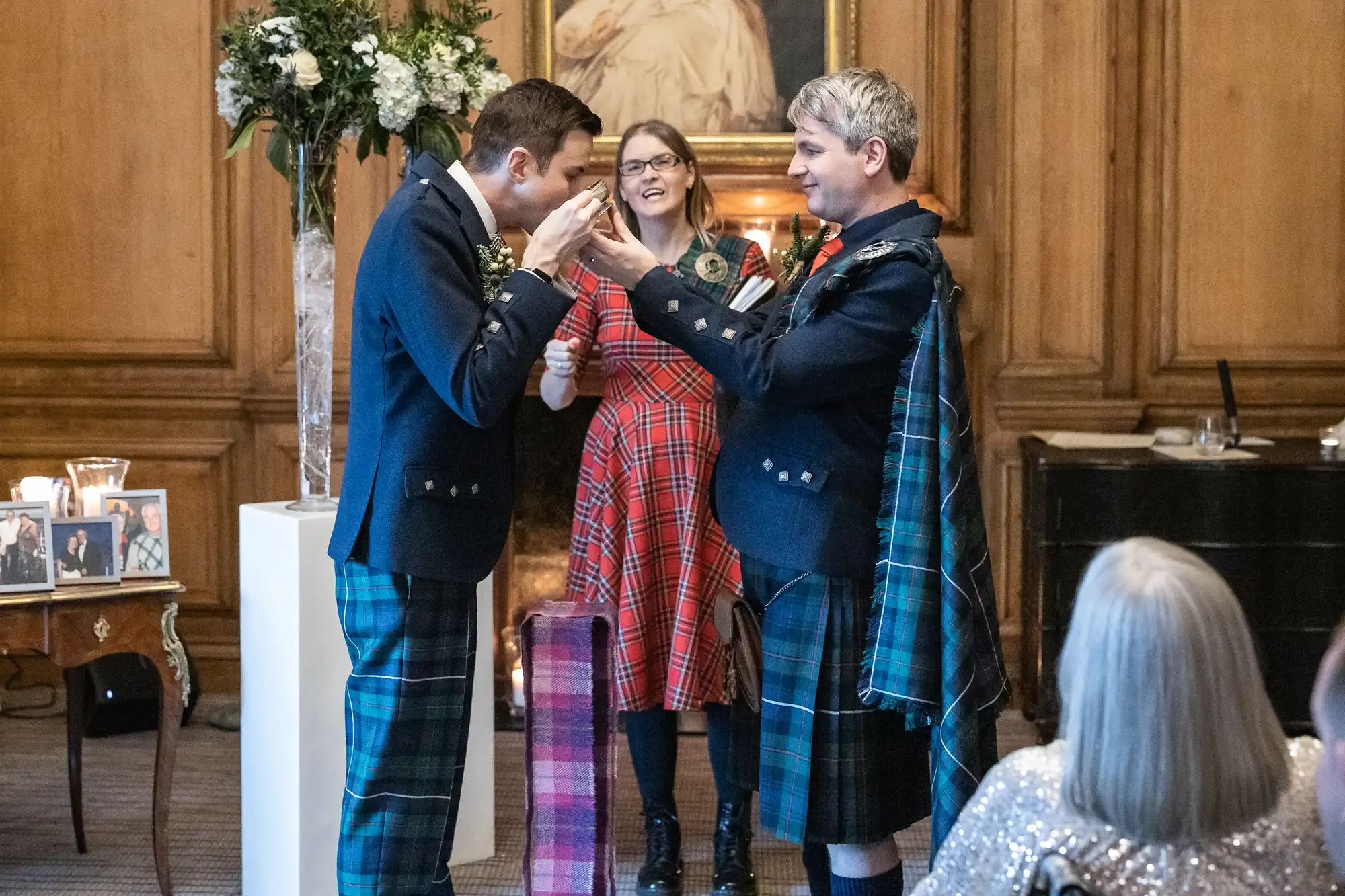 Two grooms in kilts exchange vows in front of a smiling officiant in a wood-paneled room. A woman in a gray sweater watches from the foreground.