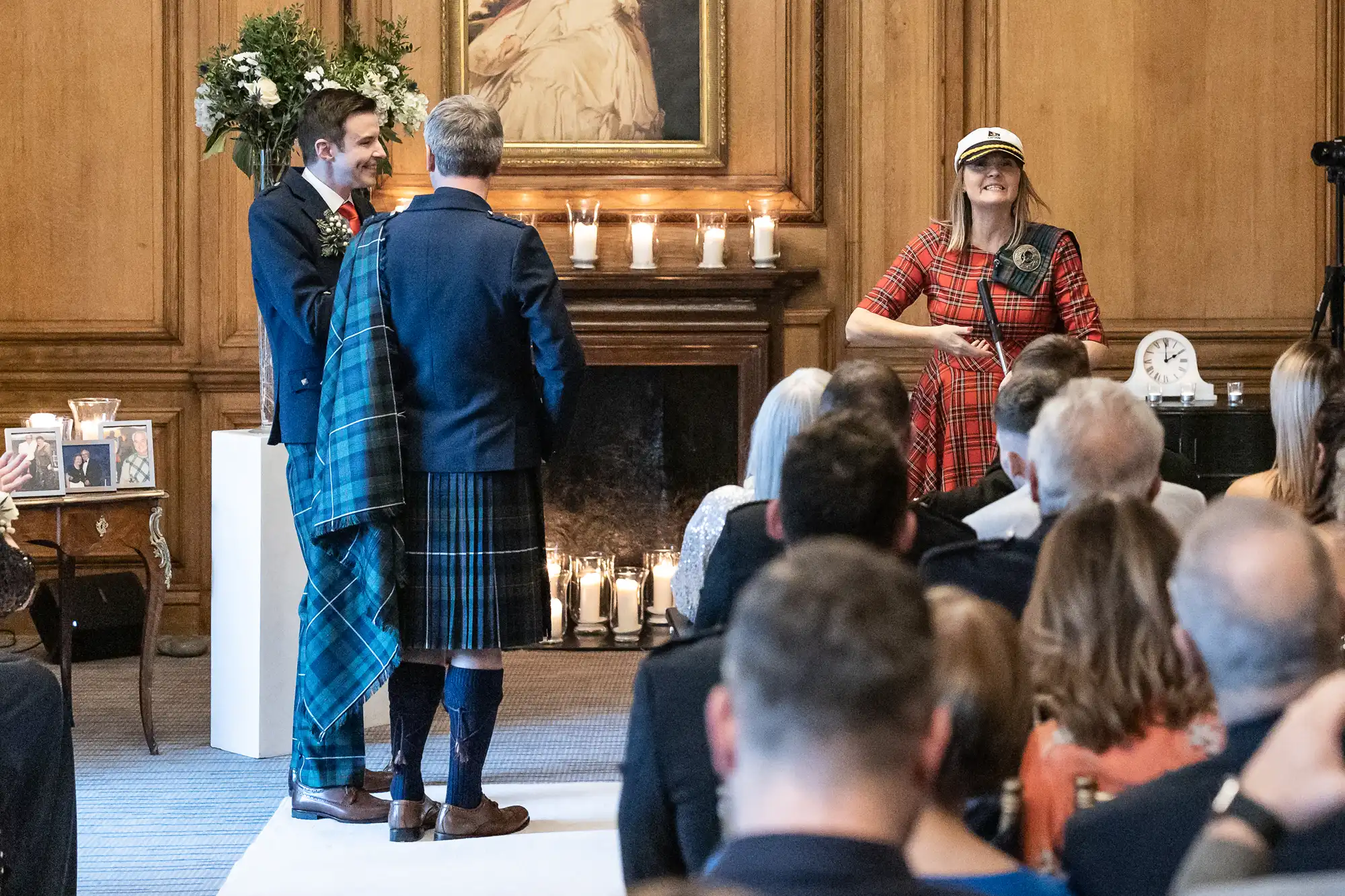 A wedding officiant in a tartan dress and cap stands before a couple, one in a kilt and the other in a suit, as they face each other during their ceremony in a wood-paneled room.