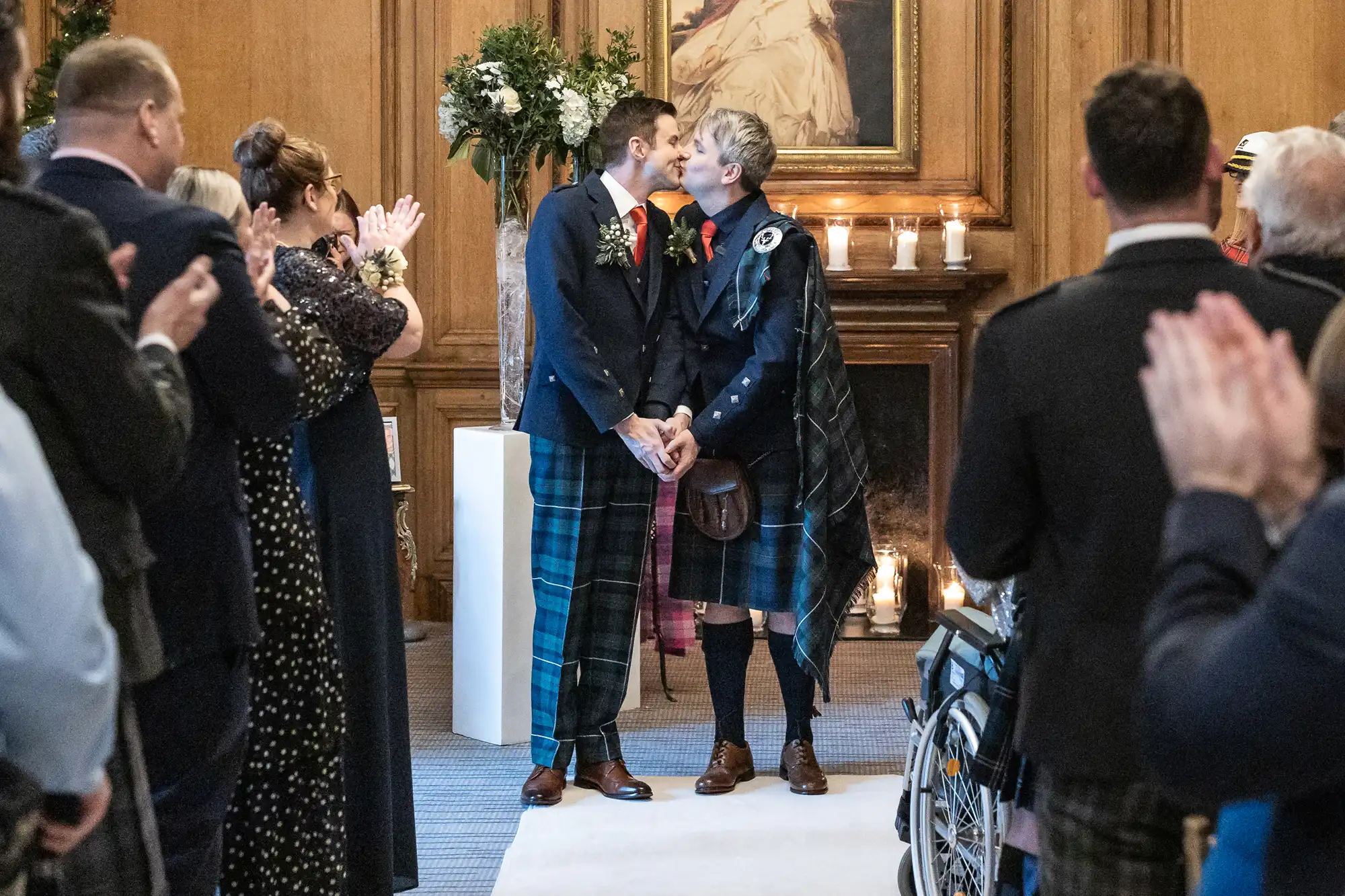 Two men in kilts kiss at the altar during their wedding ceremony, surrounded by applauding guests.