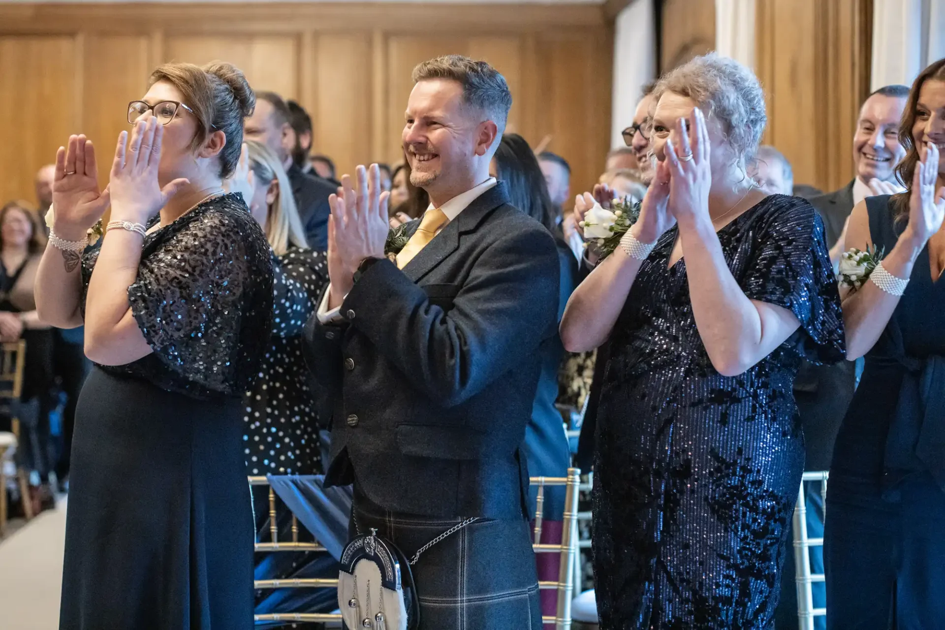 A group of people dressed in formal attire, standing and clapping, with smiles and expressions of joy, inside a room with wooden panelling during an unplugged wedding ceremony.