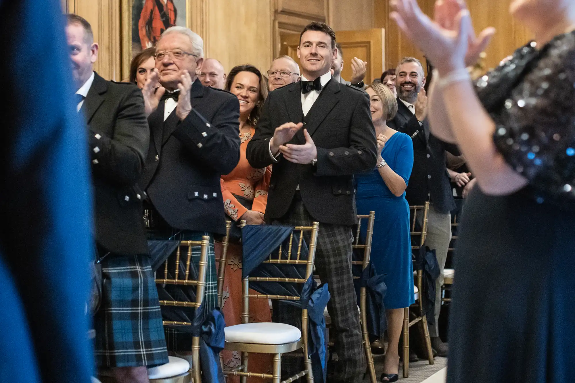 A group of people, some wearing formal attire and kilts, stand and clap in a warmly lit room with wooden walls and elegant chairs.