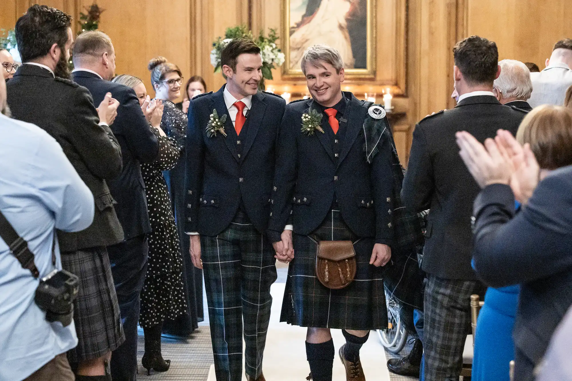 Two men in matching attire, including kilts and jackets, hold hands and smile as they walk down an aisle, surrounded by clapping guests in a warmly lit indoor setting.