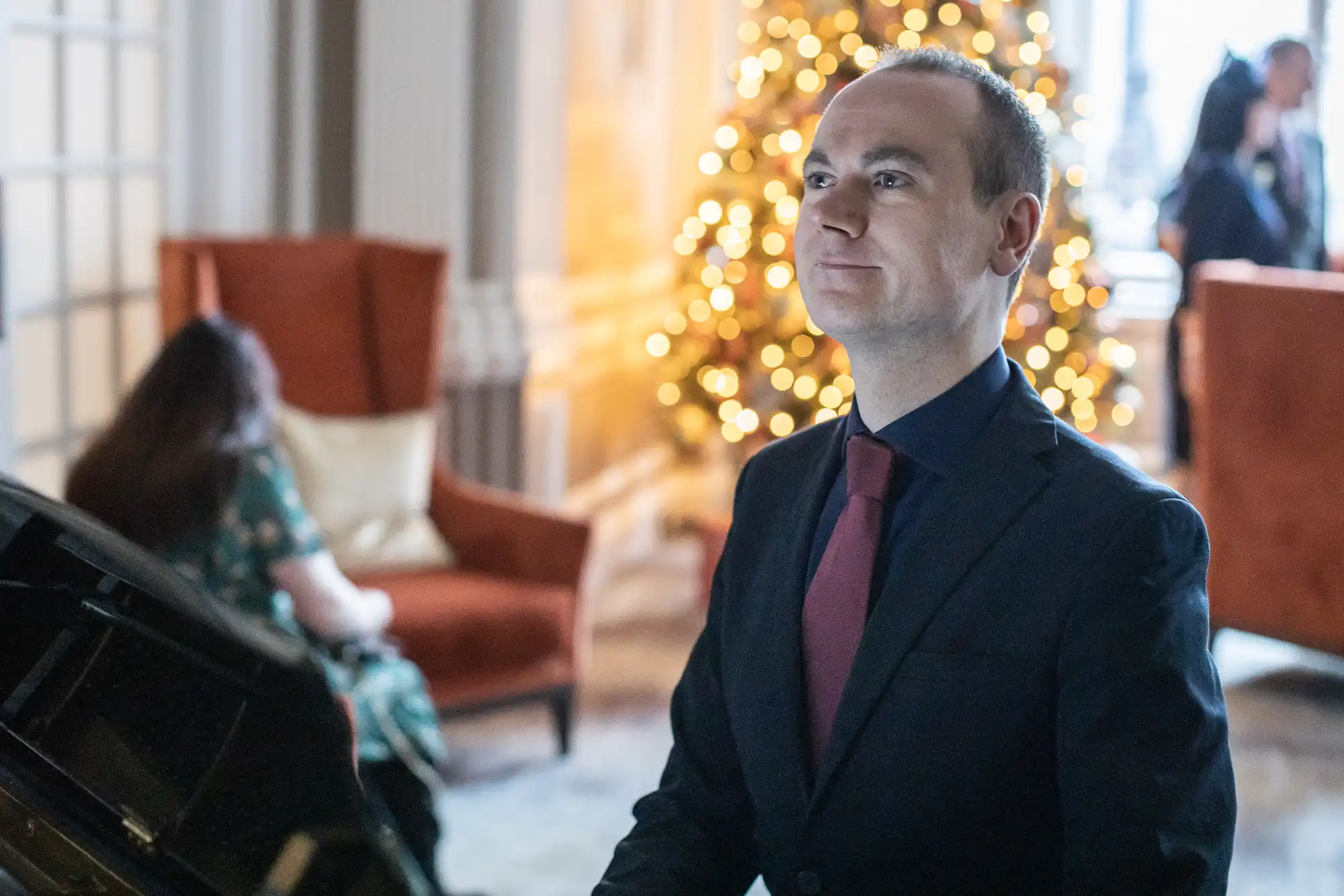 A man in formal attire stands indoors near a grand piano with a decorated Christmas tree in the background.