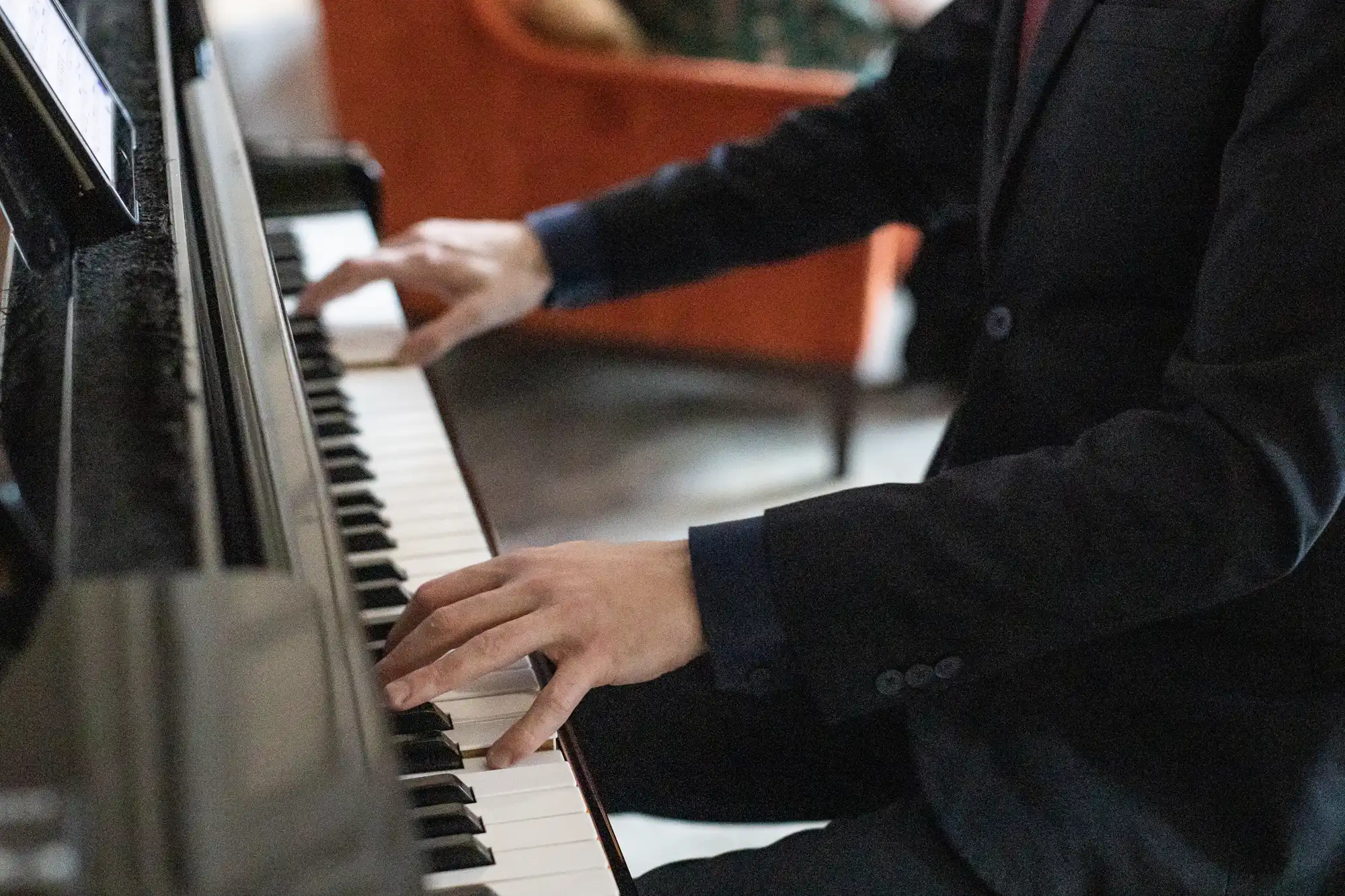 A person wearing a dark suit is playing a piano, with both hands positioned on the keys. An orange couch is visible in the background.