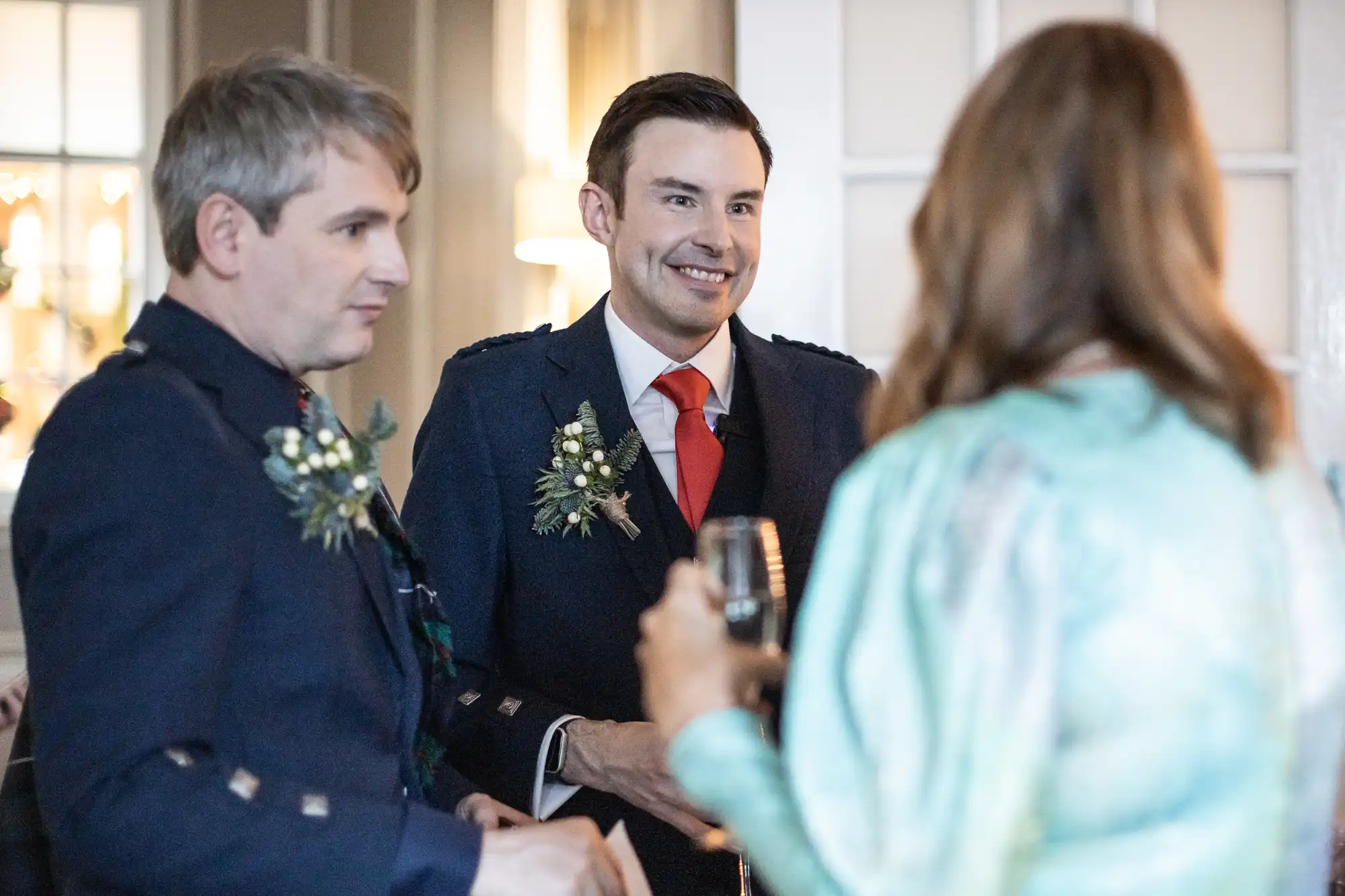 Two men in suits with floral boutonnieres are in conversation with a woman holding a drink at a social event.