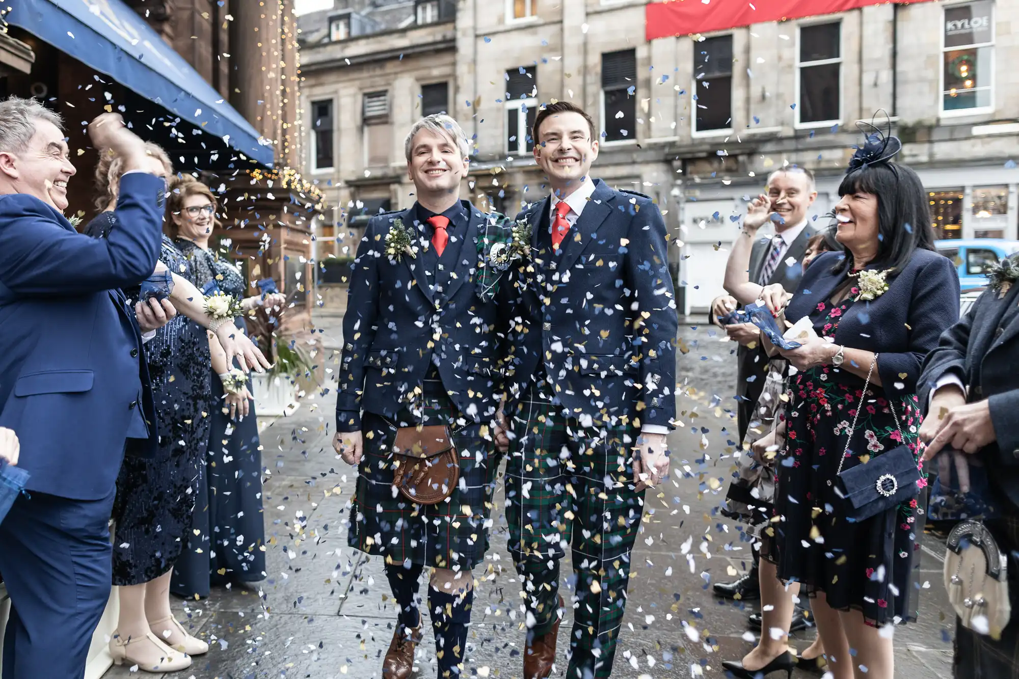 Two men, dressed in kilts and suits, walk down a street while surrounded by people throwing confetti. The background features a building and onlookers dressed in formal attire.