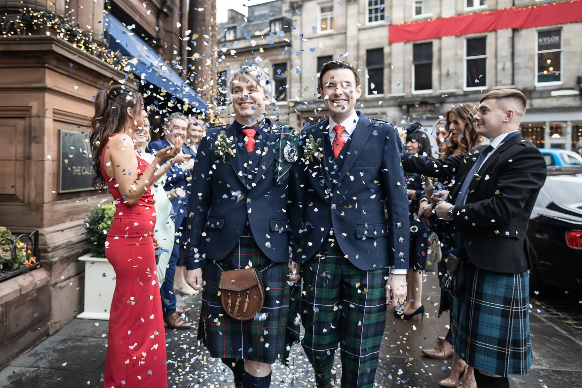 Two men in kilts walk hand in hand under a shower of confetti, surrounded by smiling guests dressed in formal attire. A woman in a red dress stands nearby. They appear to be celebrating a special occasion.