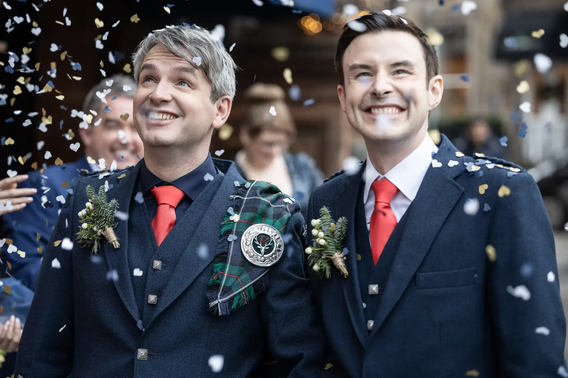 Two grooms in matching blue suits with red ties smile as they are showered with confetti. Both have boutonnières, one has a plaid draped over his shoulder. They look joyful.