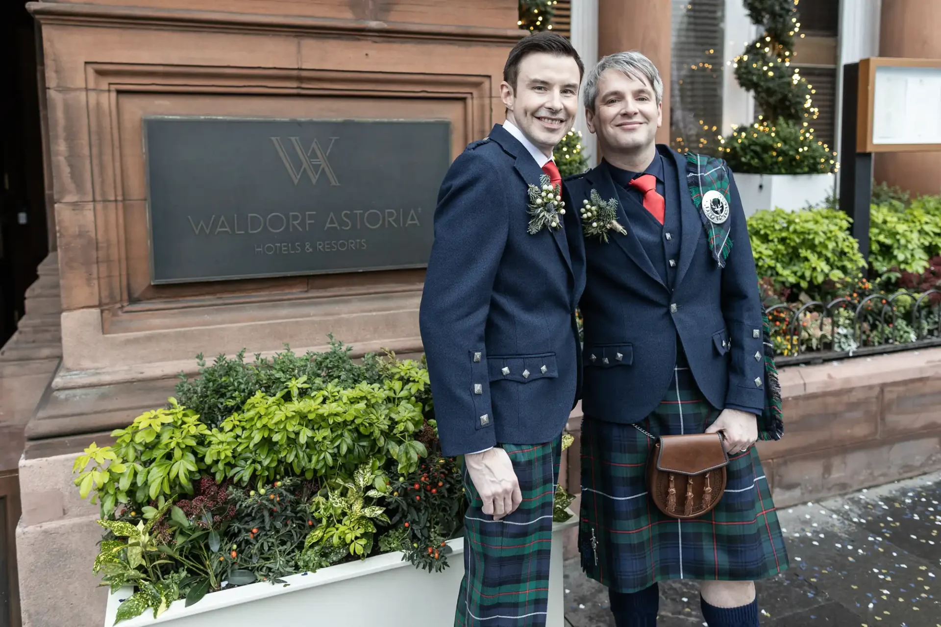Two men wearing traditional Scottish kilts stand side by side smiling outside the Waldorf Astoria hotel, with festive decorations in the background.