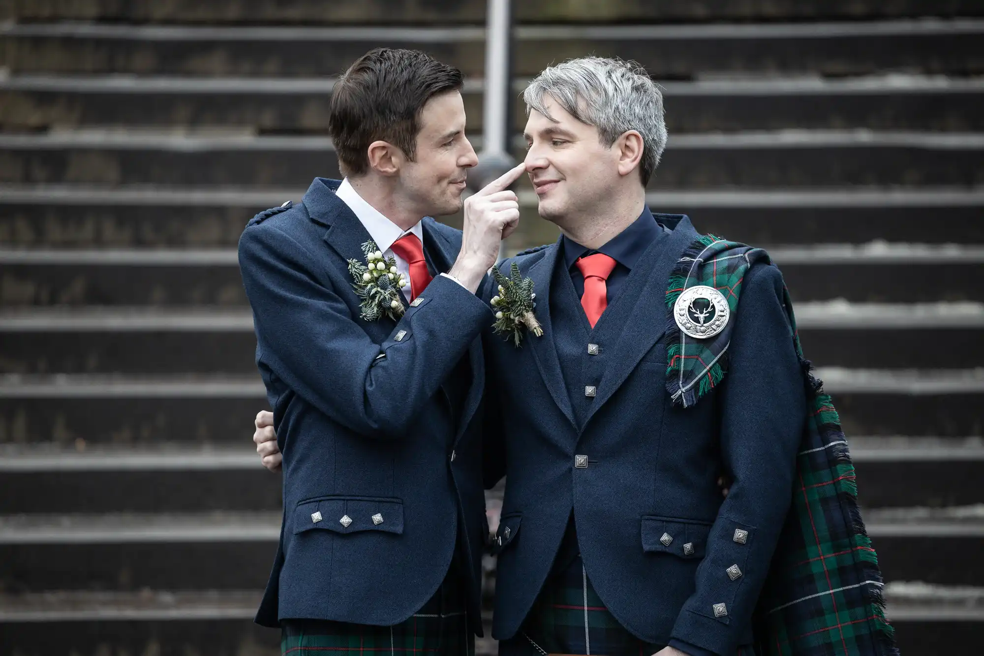 Two men in matching formal attire, including kilts and red ties, stand before steps. One man gestures affectionately to the other, who wears a tartan sash over his shoulder. Both are smiling.