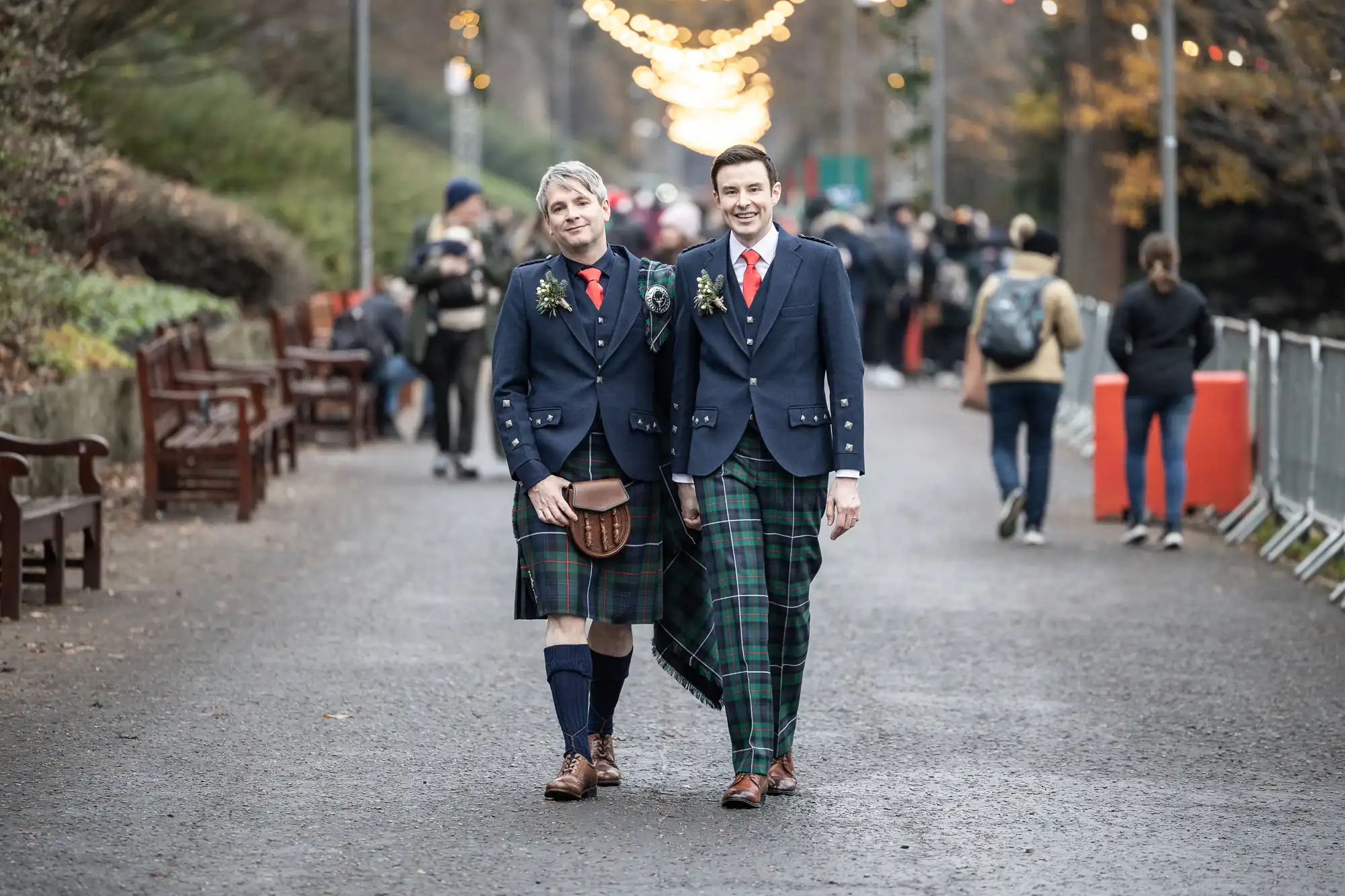 Two individuals dressed in Scottish kilts and formal jackets walk down a street lined with people and trees decorated with string lights.