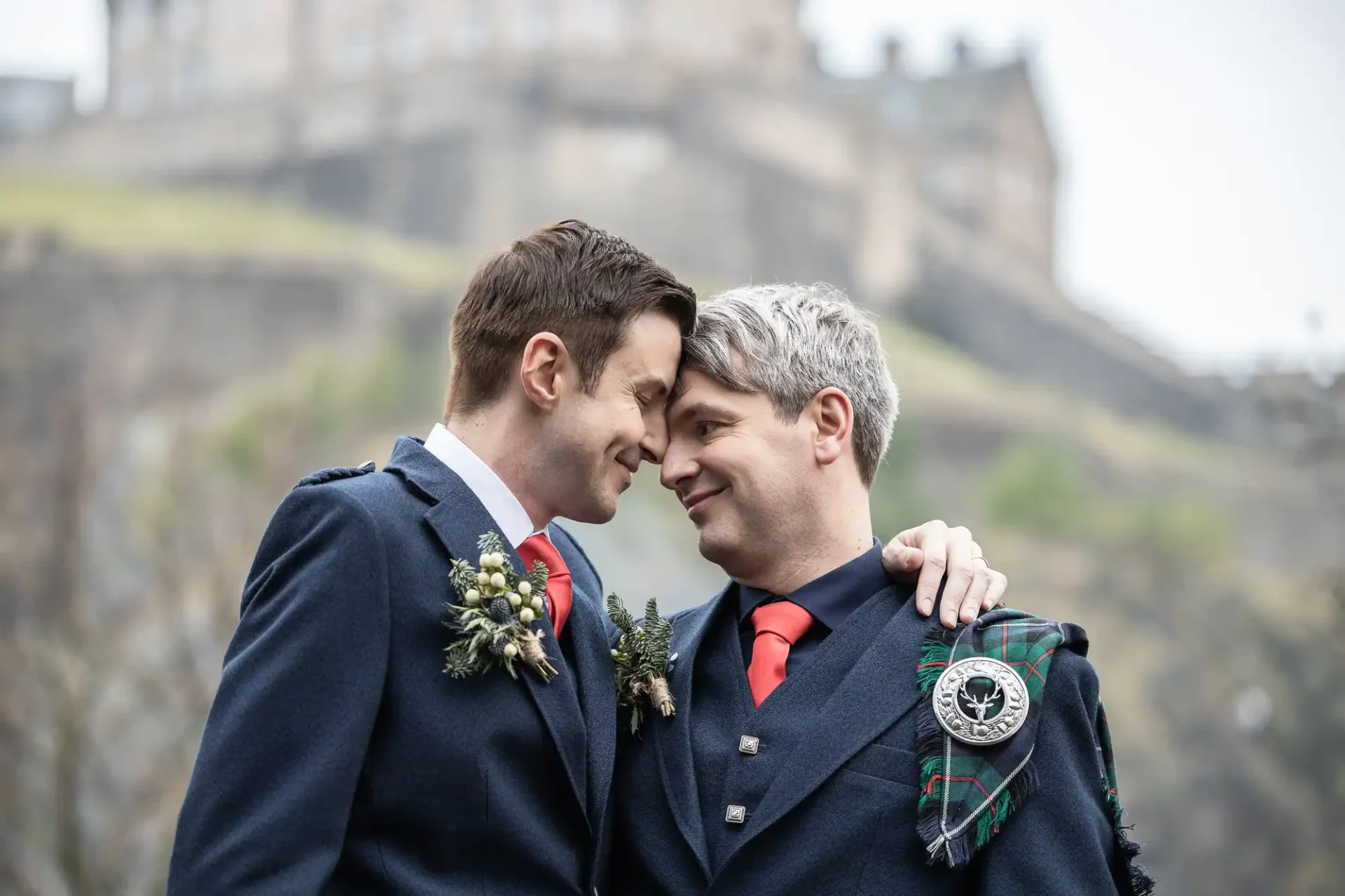 Caledonian Hotel wedding photographers: Two men in traditional Scottish attire share an affectionate moment outdoors, with one wrapping his arm around the other. They stand close, smiling at each other. A historic building is in the background.