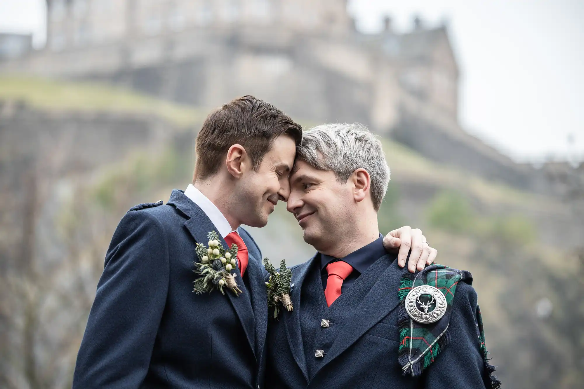 Two men in traditional Scottish attire share an affectionate moment outdoors, with one wrapping his arm around the other. They stand close, smiling at each other. A historic building is in the background.