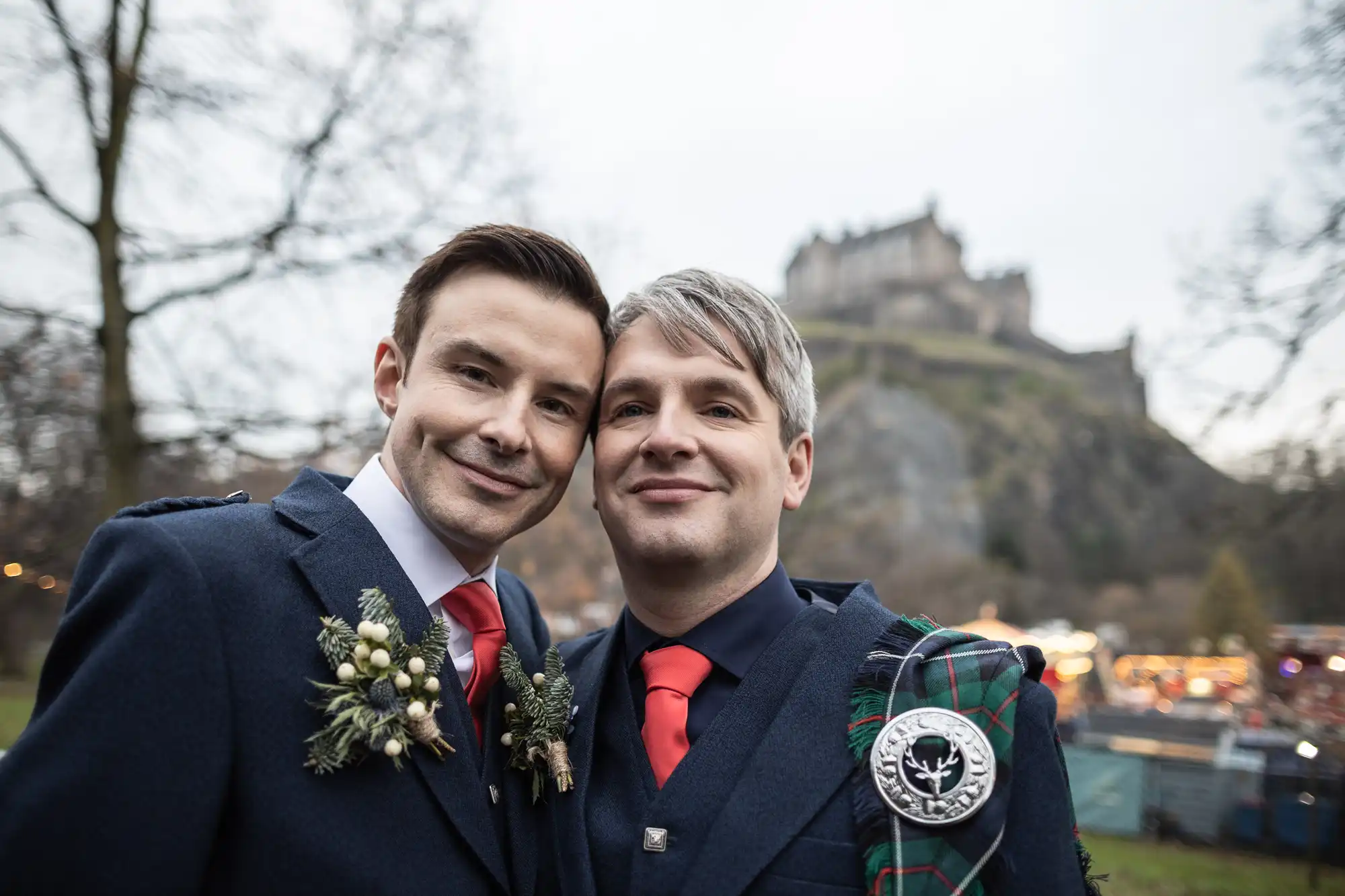Two men in formal attire and red ties stand closely together, smiling, at an outdoor location with a large castle in the background.