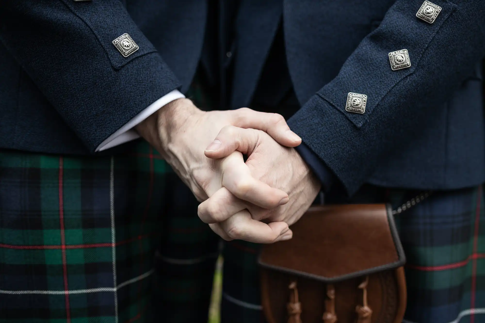 Two people in tartan kilts stand close together, with their hands clasped. They are wearing dark jackets with metal buttons and a small leather pouch.