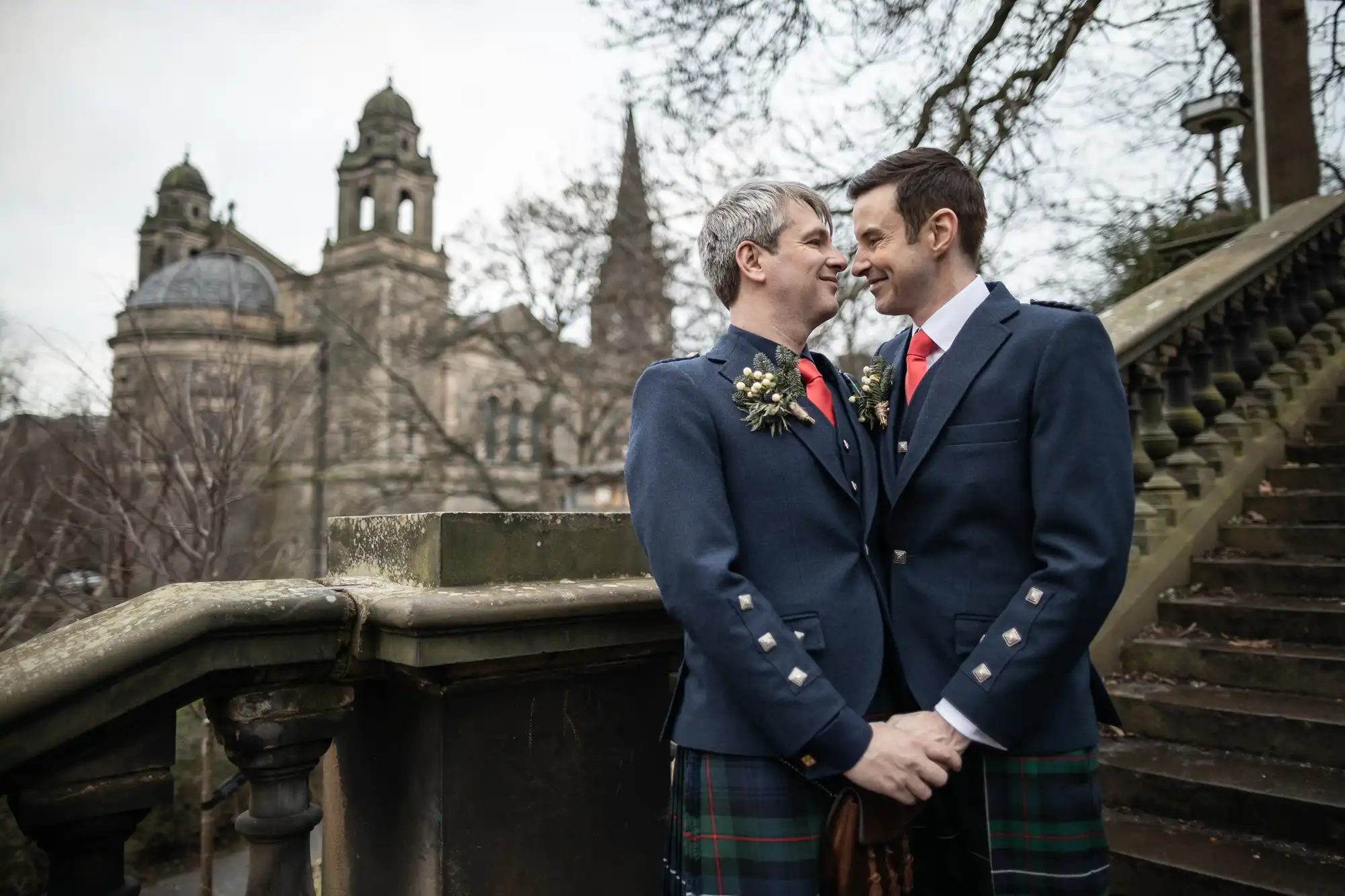 Two men in matching formal attire hold hands and gaze at each other lovingly on an outdoor staircase, with historical architecture and bare trees in the background.