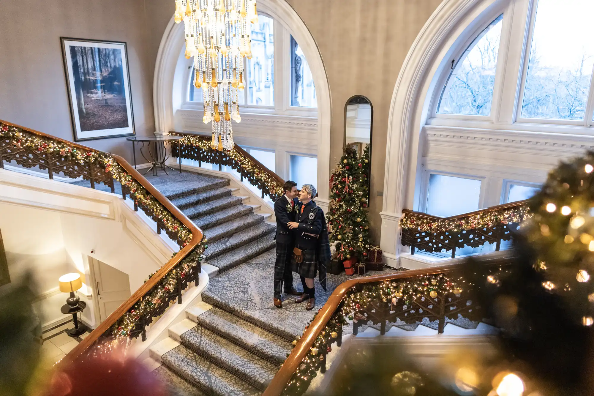Two people in formal attire hold hands and smile at each other on a grand staircase decorated with holiday lights and garlands in an elegant building with arched windows and a large chandelier.