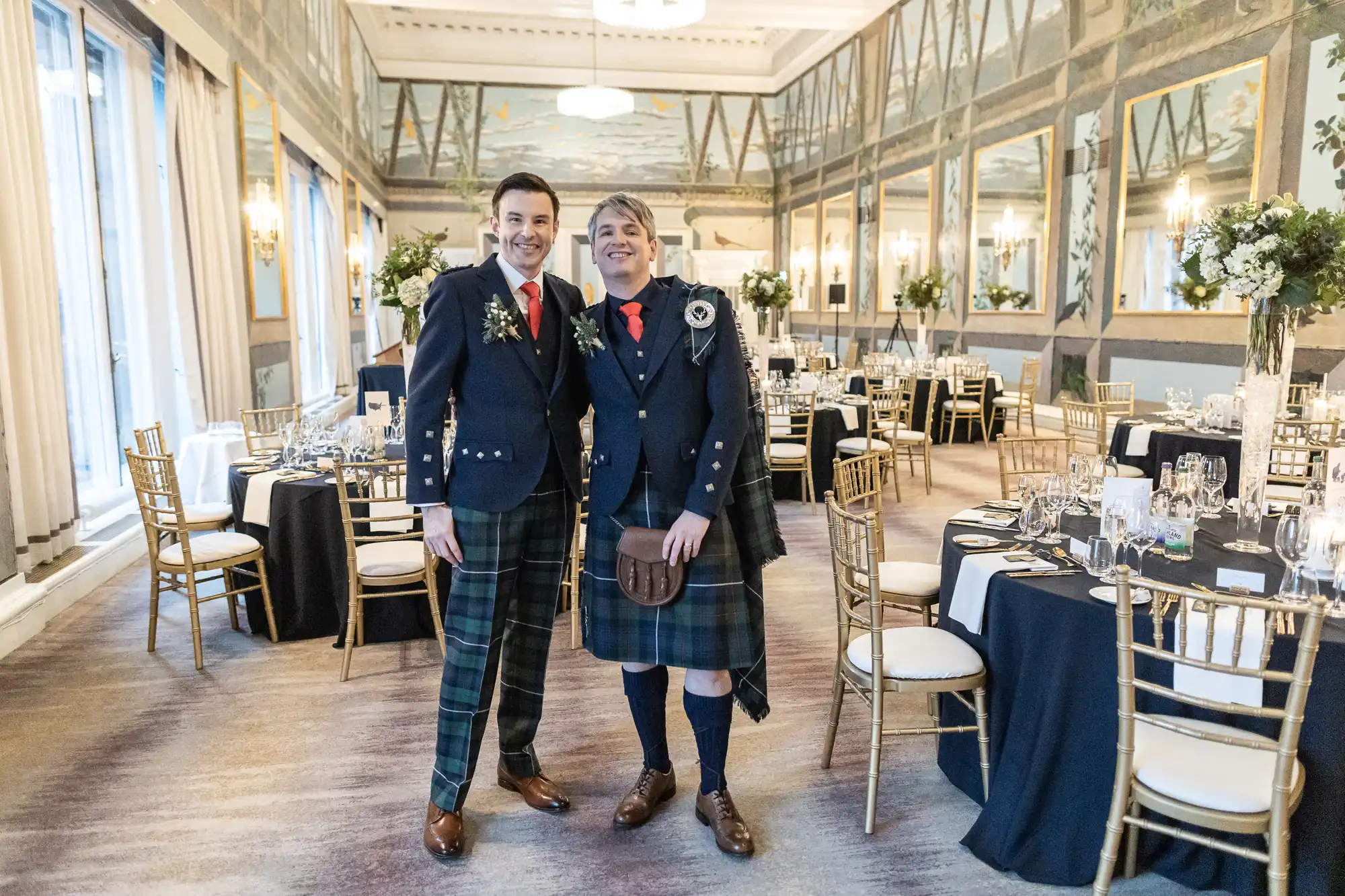 Two men in traditional Scottish kilts stand smiling in an elegantly decorated banquet hall with set tables and gold chiavari chairs.