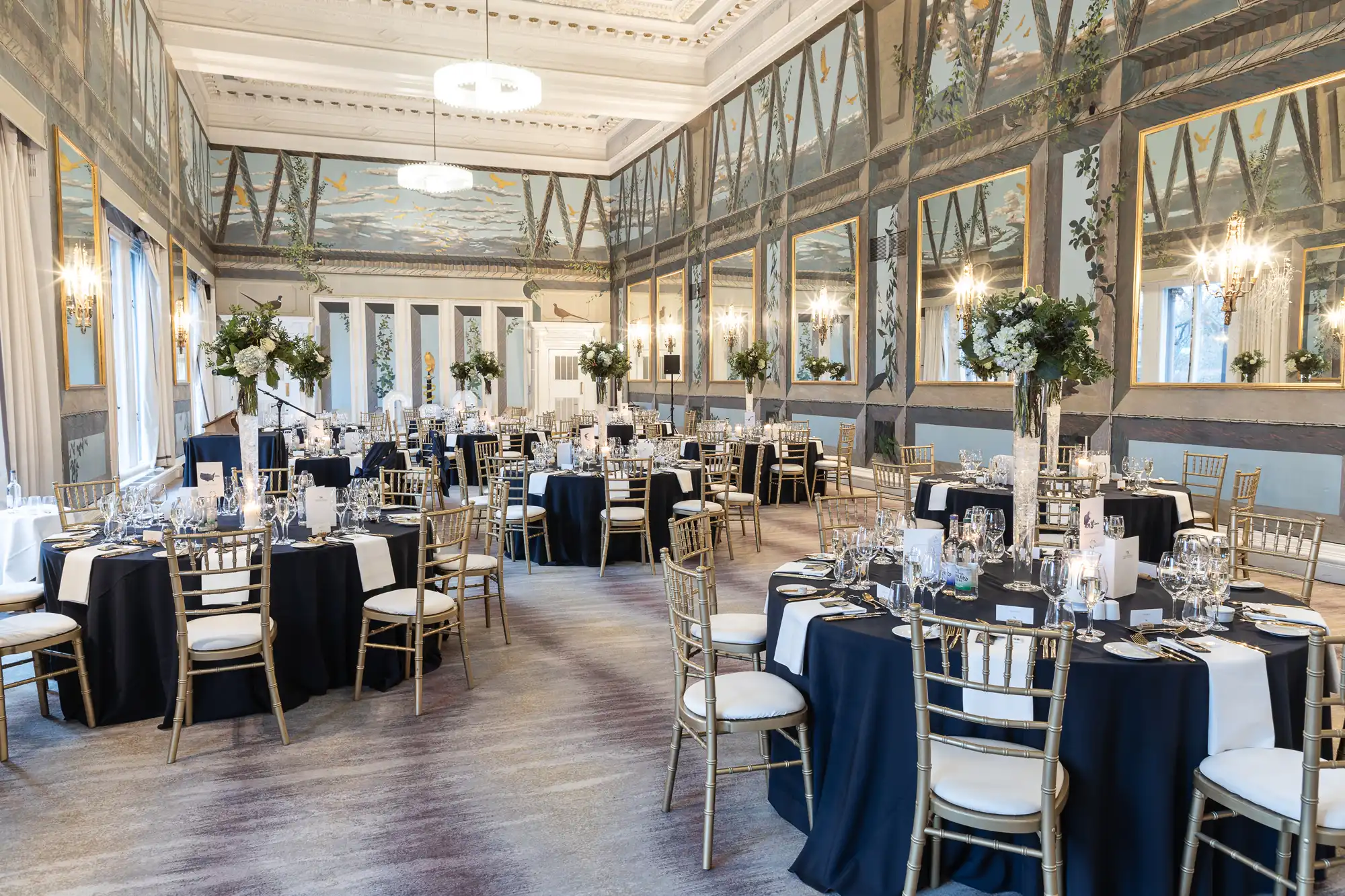 A formal dining room with elegantly set tables, white and black tablecloths, tall floral centerpieces, and gold chairs is prepared for an event. The walls are adorned with large mirrors and detailed murals.