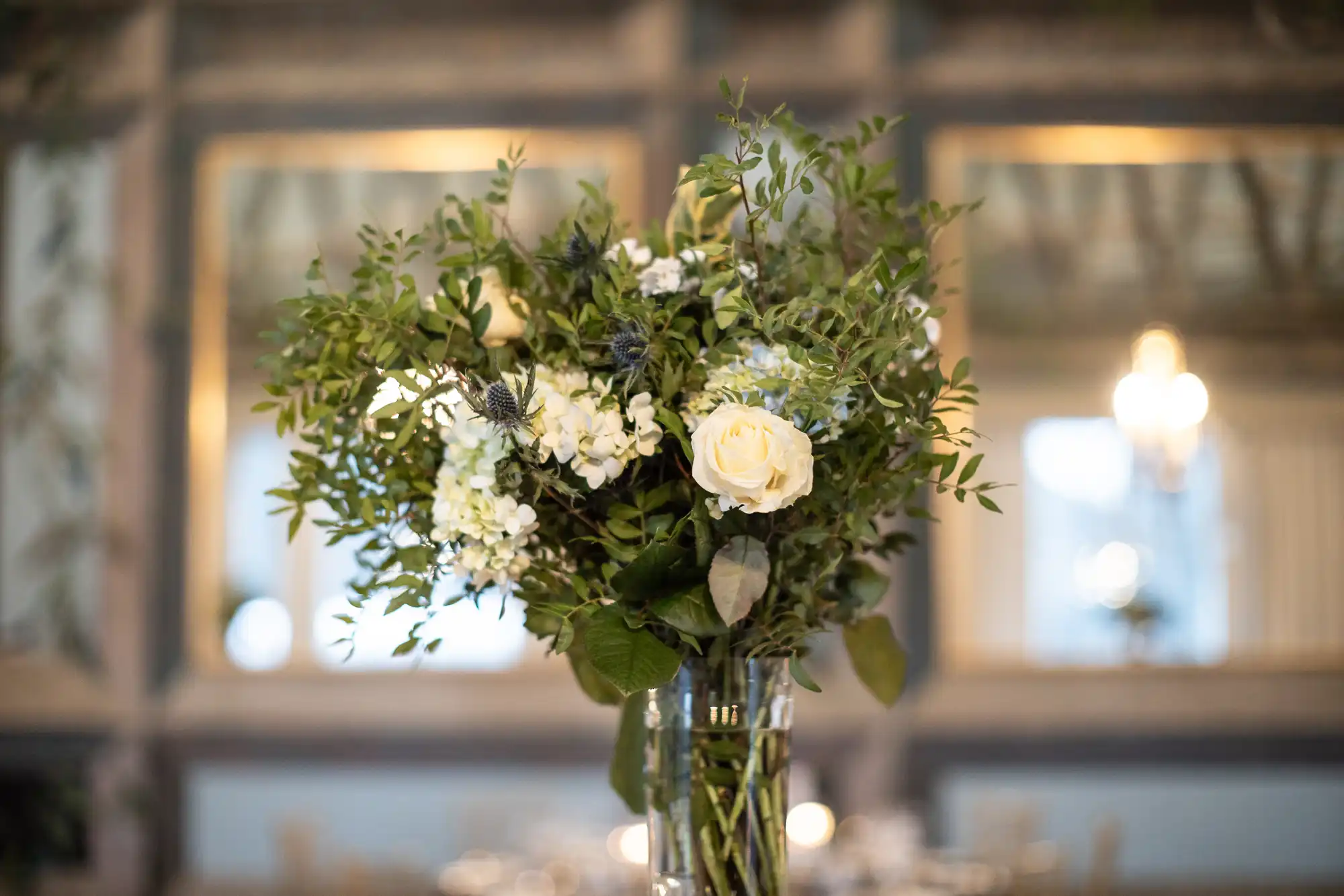 A vase with a mixed floral bouquet, including white roses and greenery, is placed on a table. There are mirrors and blurred lights in the background.