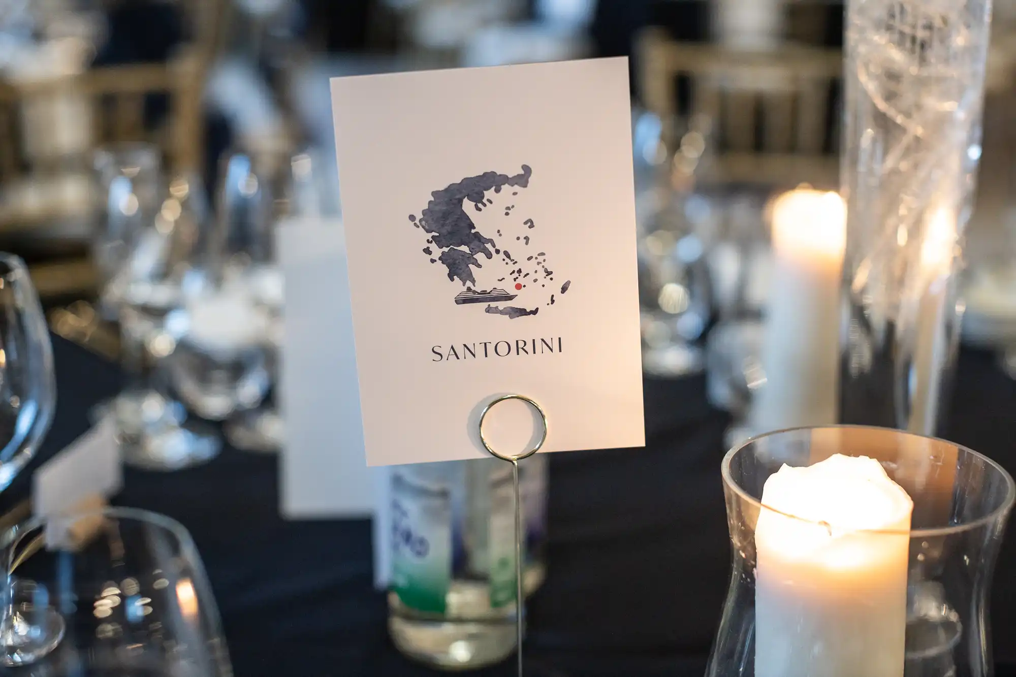 A table card labeled "Santorini" with a map illustration is placed in a holder on a well-set dining table with glasses, candles, and a decorative centerpiece.