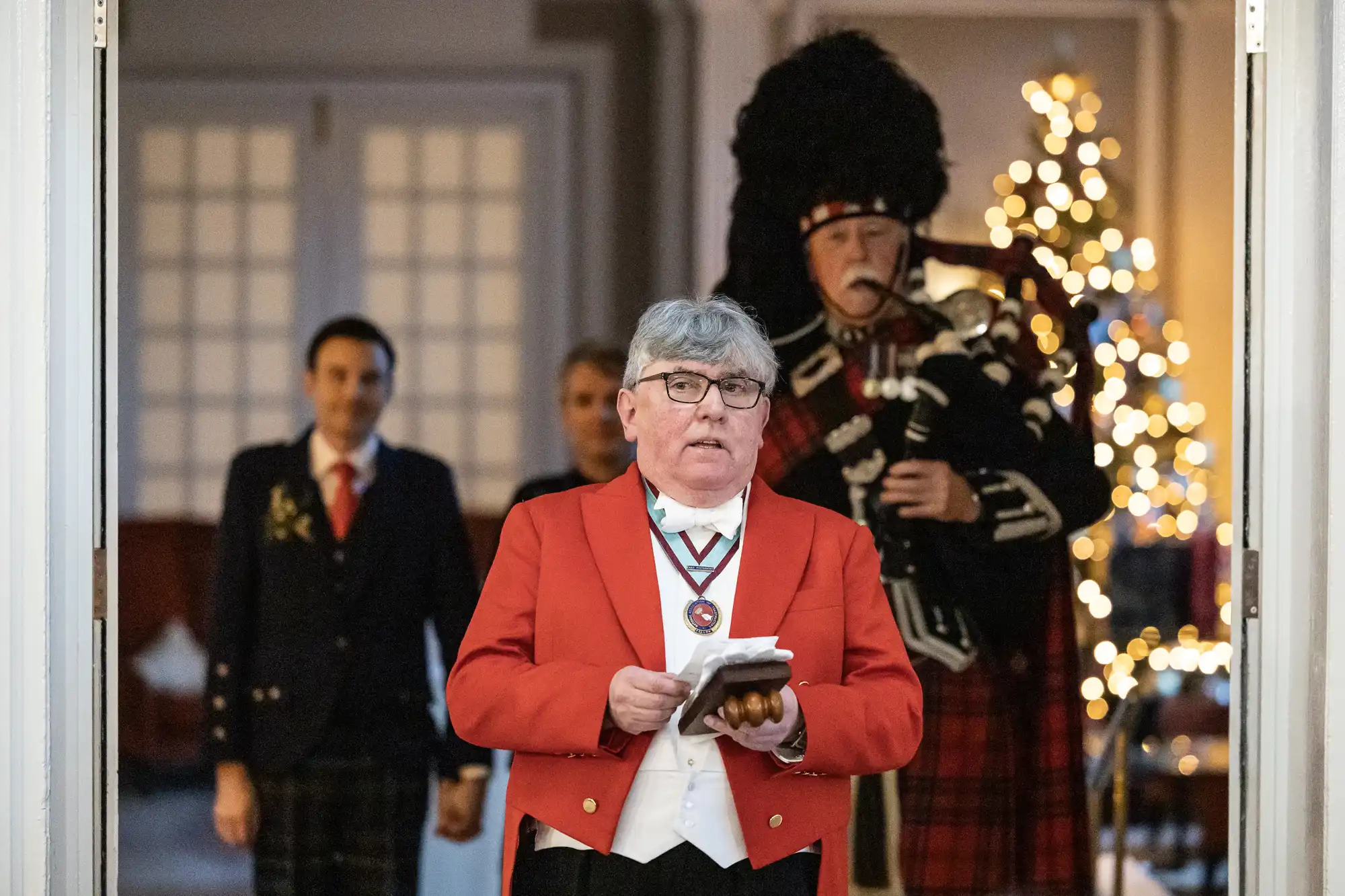 A man in a red jacket speaks while holding a notepad, flanked by people in formal attire. A man in traditional Scottish attire and a bagpipe stands behind him. A decorated Christmas tree is visible.