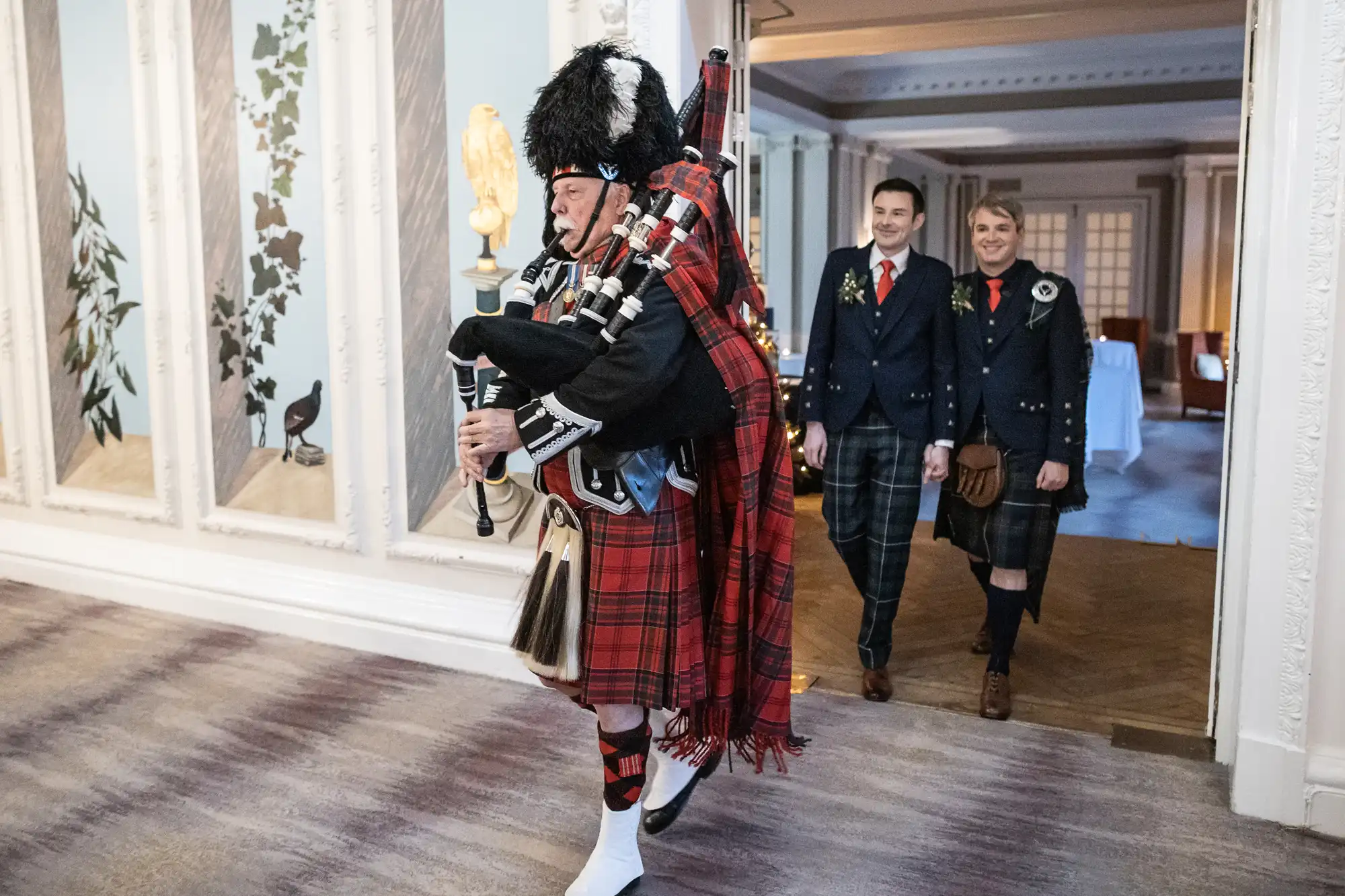 A person dressed in traditional Scottish attire plays bagpipes while walking, followed by two people in similar attire. They are indoors in an ornate room with white walls and wood flooring.