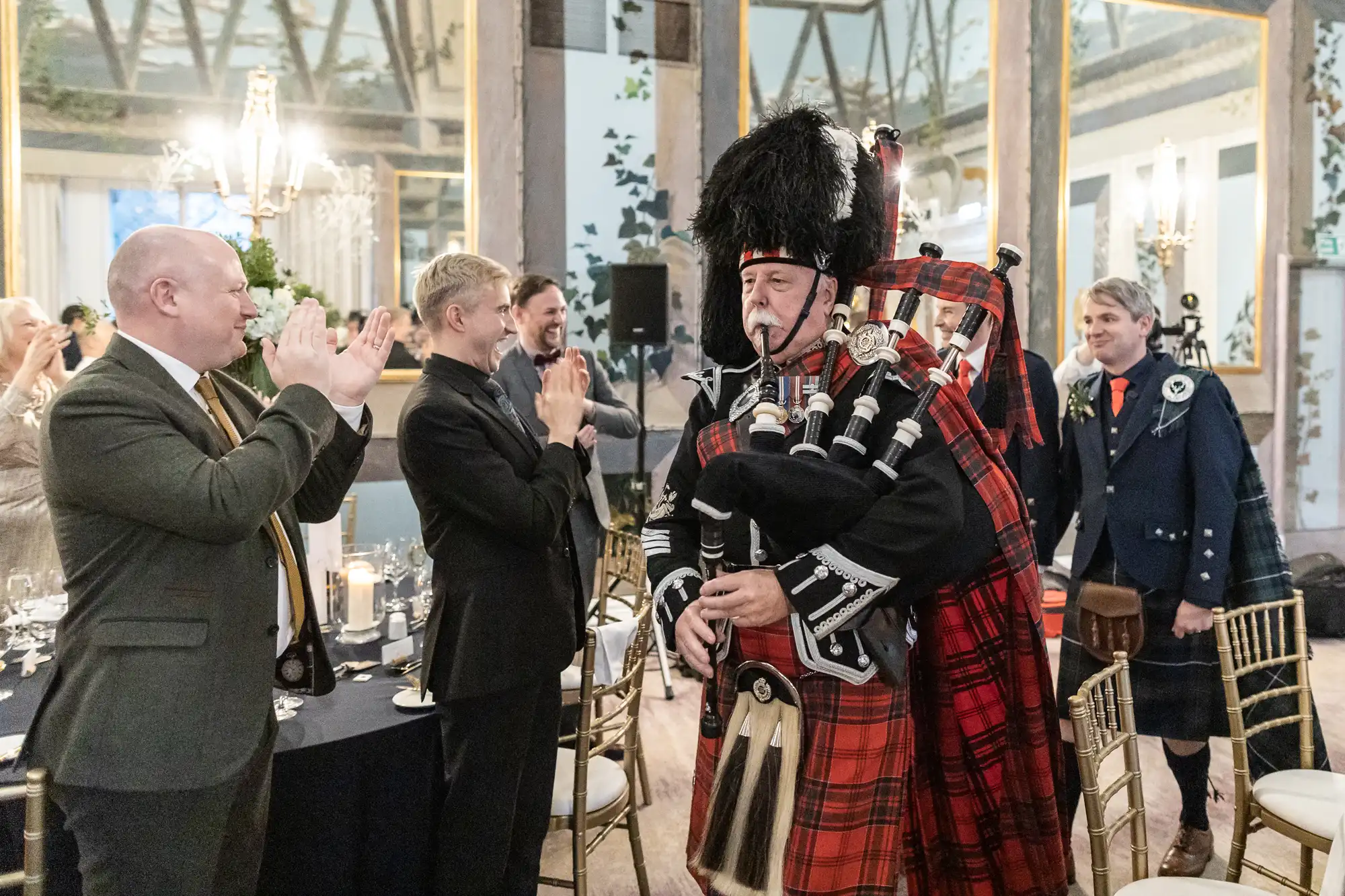 A bagpiper in traditional Scottish attire plays as guests clap and cheer inside an elegantly decorated venue.