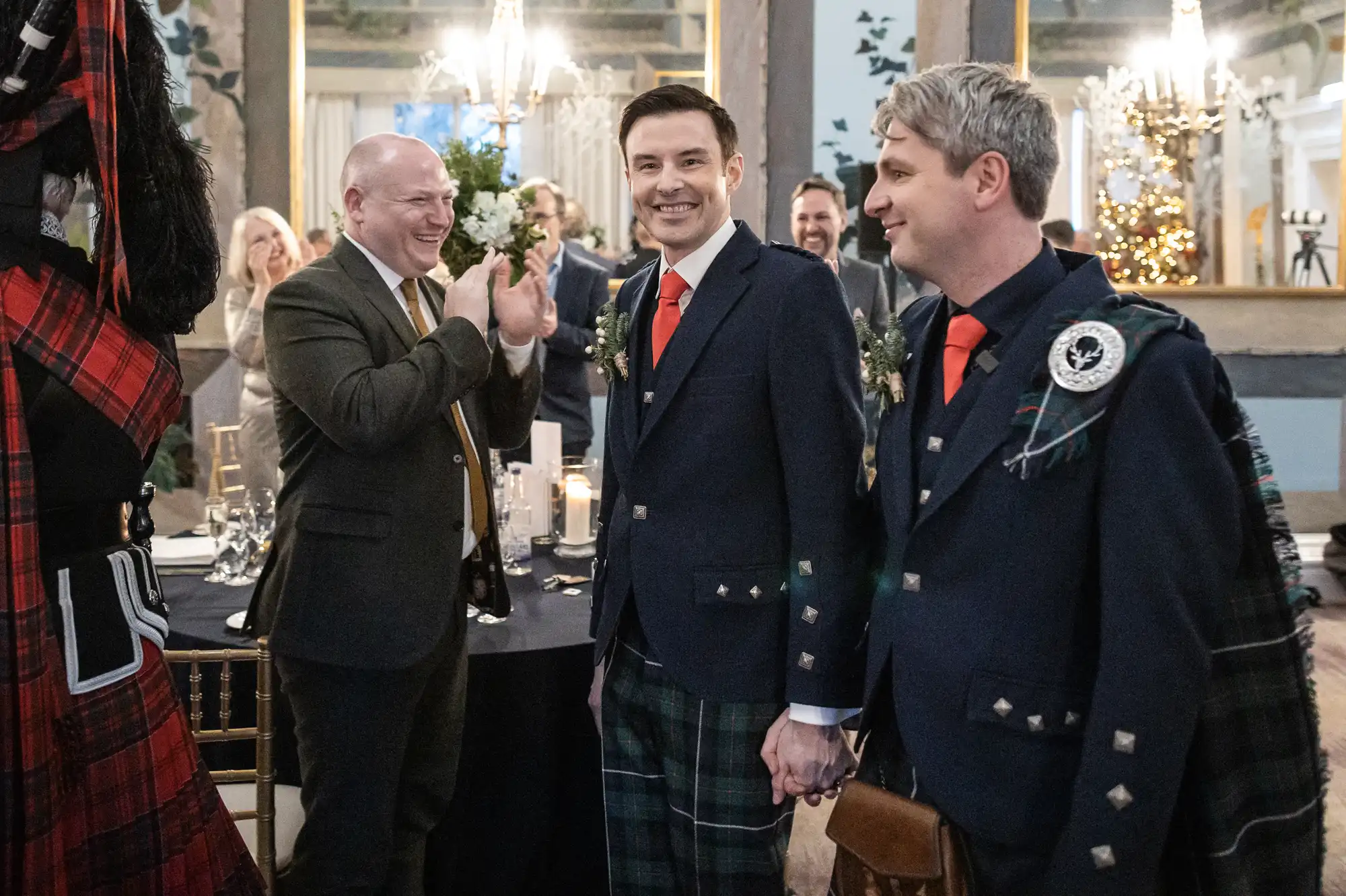 Two men in suits and kilts hold hands and smile while others around them applaud in a decorated room with tables and mirrors, suggesting a celebratory event.