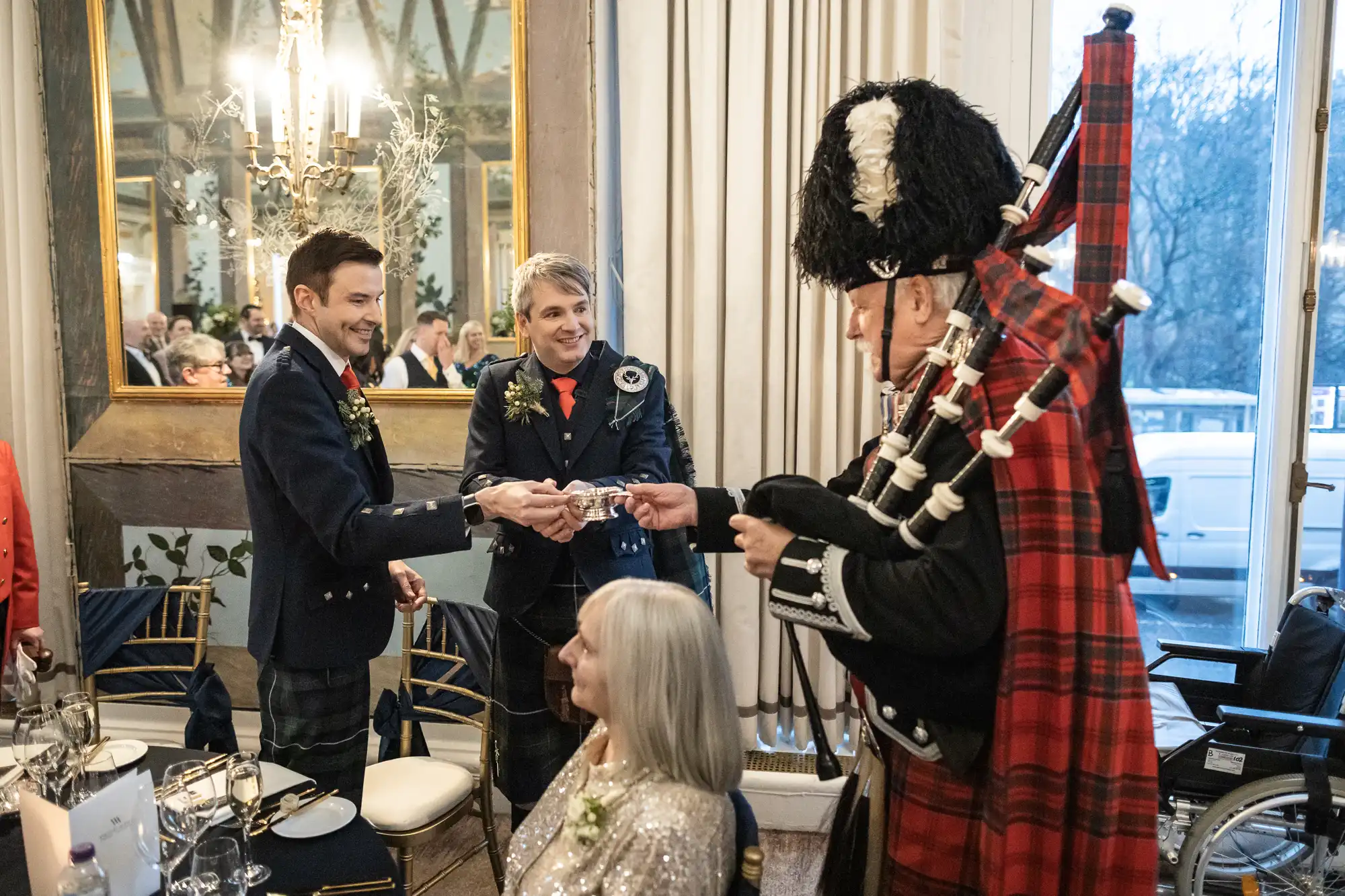 A man in traditional Scottish attire, including a kilt and feathered hat, plays bagpipes while two other men in kilts shake hands at a formal gathering. A woman with grey hair is seated nearby.