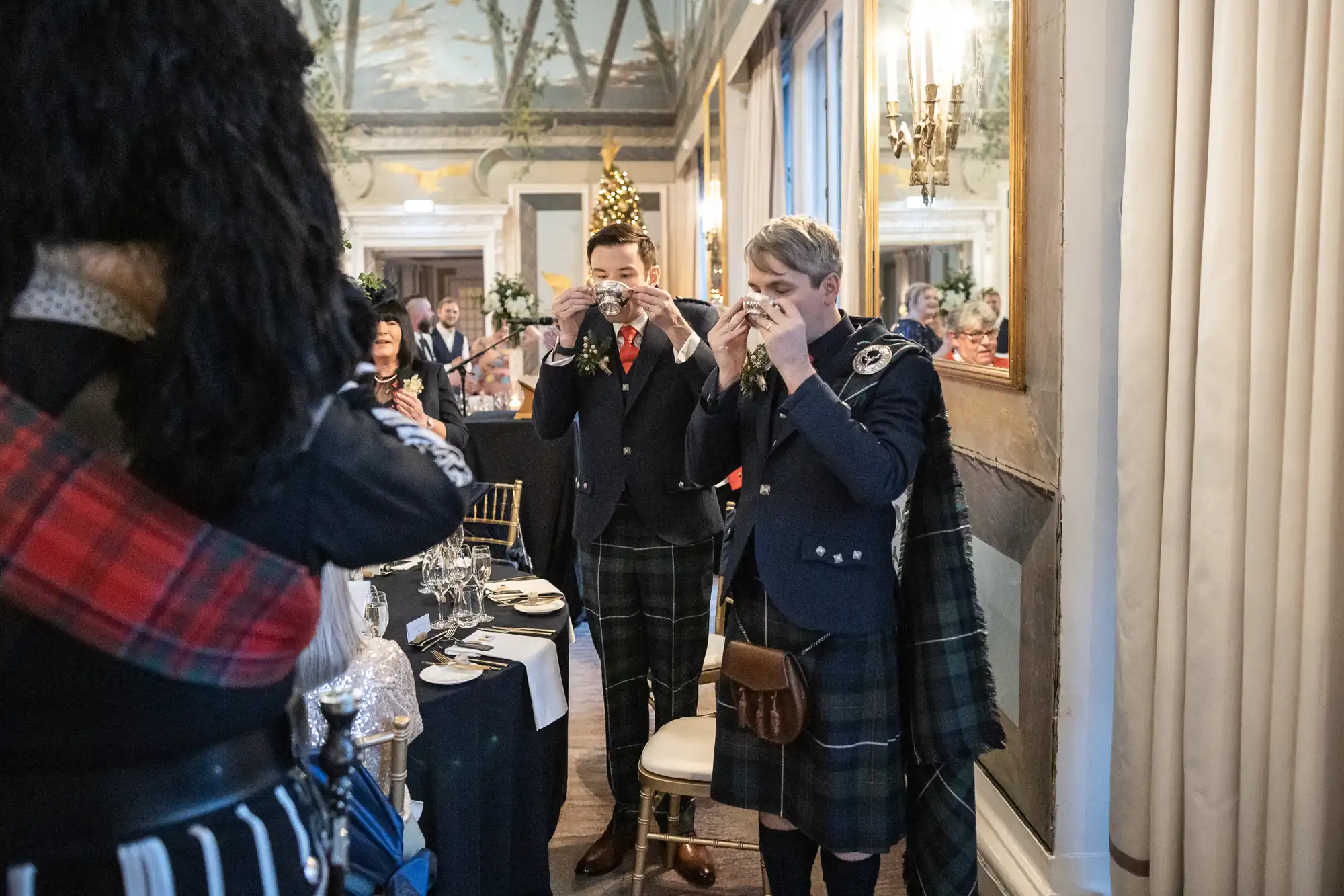 Two people in kilts and matching attire participate in a traditional toasting ceremony in an elegant room with a Christmas tree. They raise small glasses, while others look on.