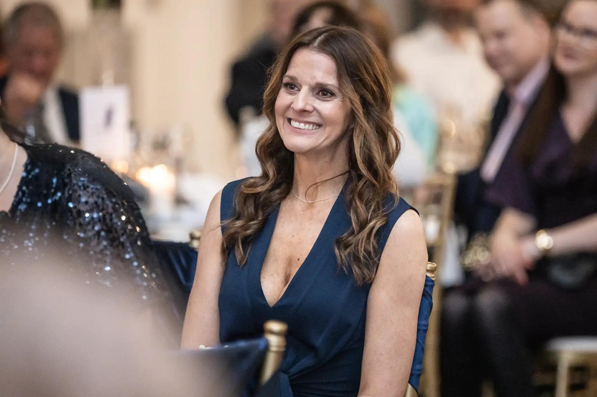 A woman with long brown hair wearing a dark blue dress smiles while seated at an indoor formal event.