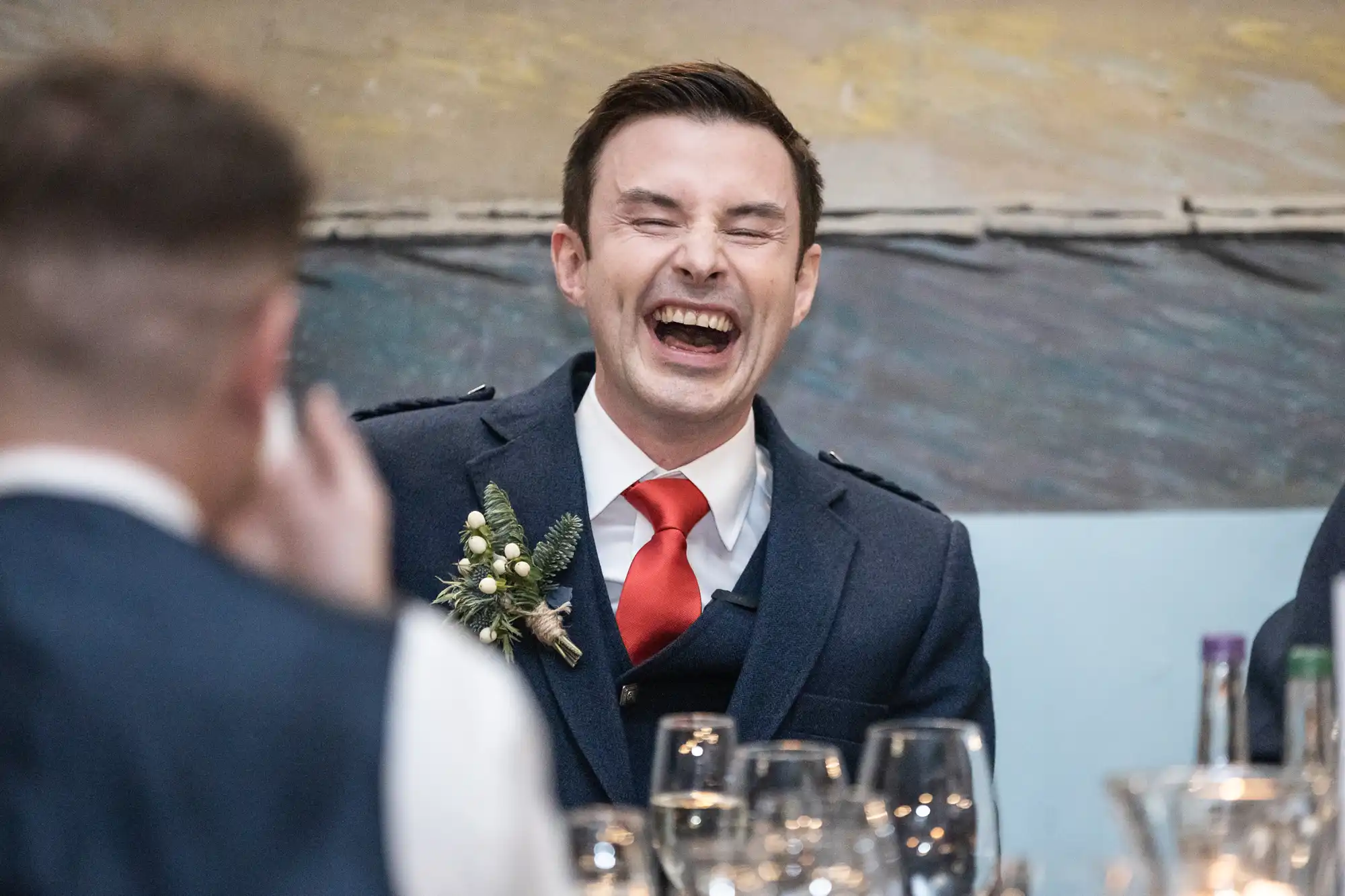 A man in a suit and red tie laughs heartily at a social event, with glassware visible on the table in front of him.