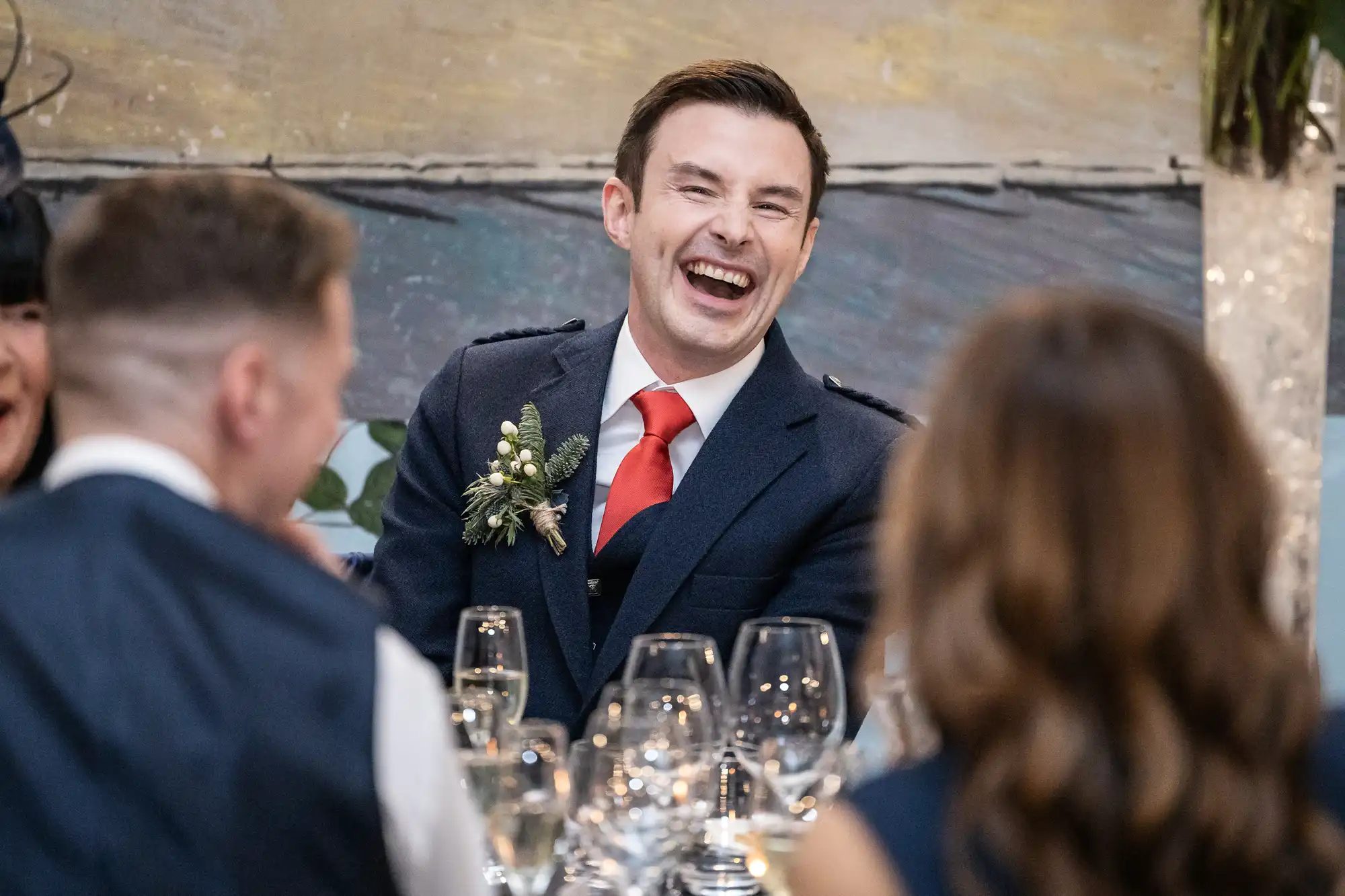 A man in a suit and red tie is laughing while sitting at a table with several people and drinking glasses in front of him.