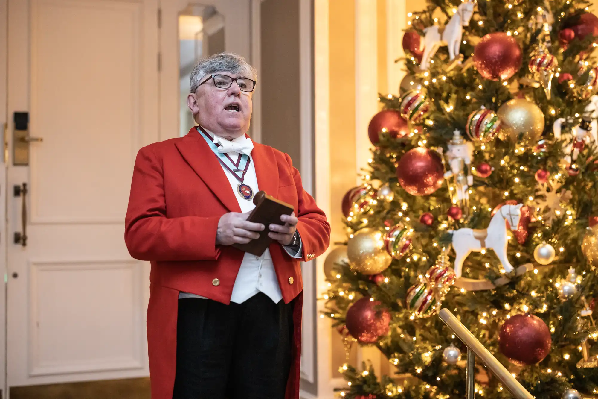 An elderly man in a red coat speaks while standing next to a Christmas tree decorated with lights, ornaments, and toy horse figures.