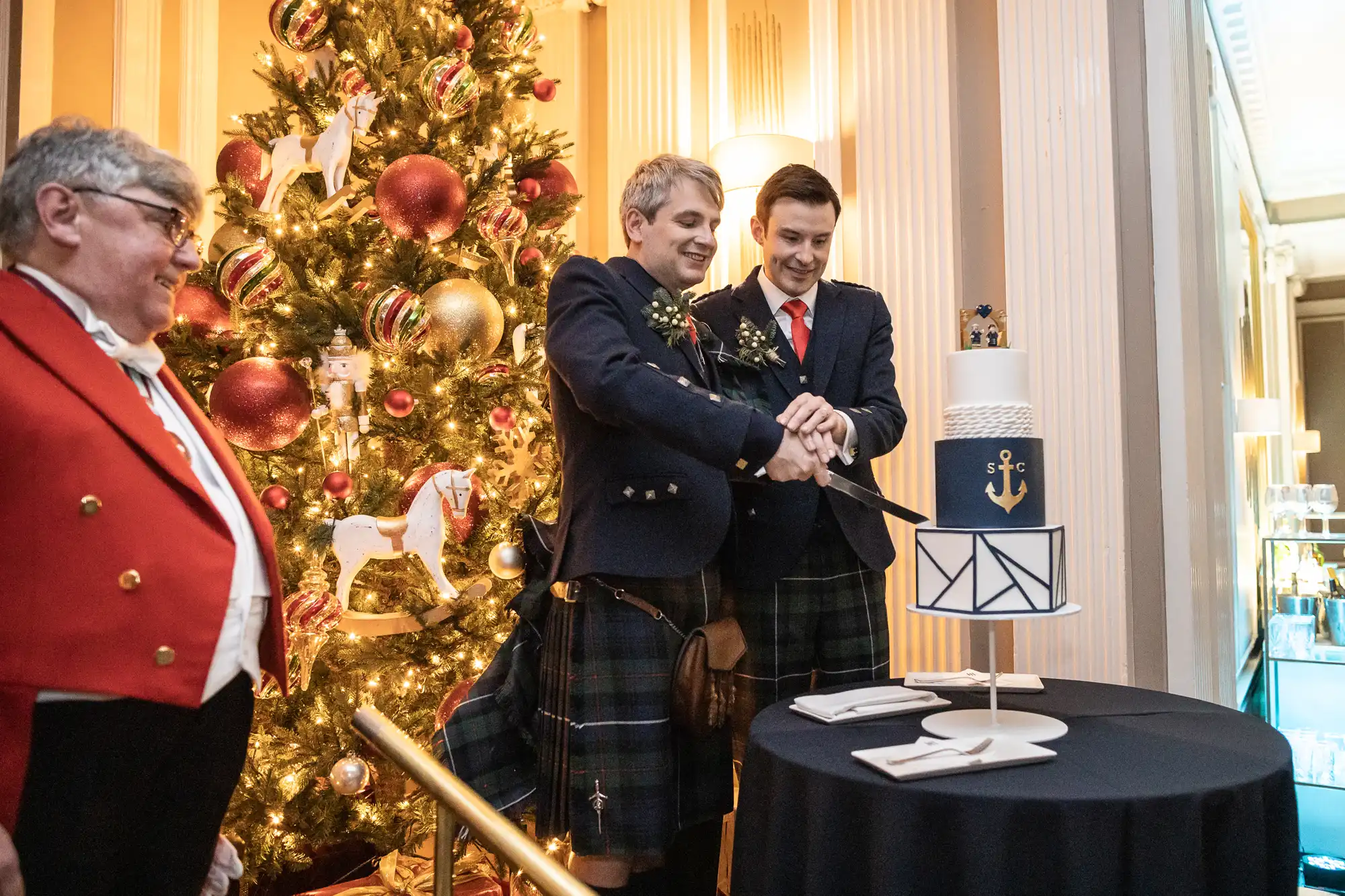 Two men in kilts are cutting a tiered cake together at a ceremony in front of a Christmas tree, while another person in a red jacket watches.
